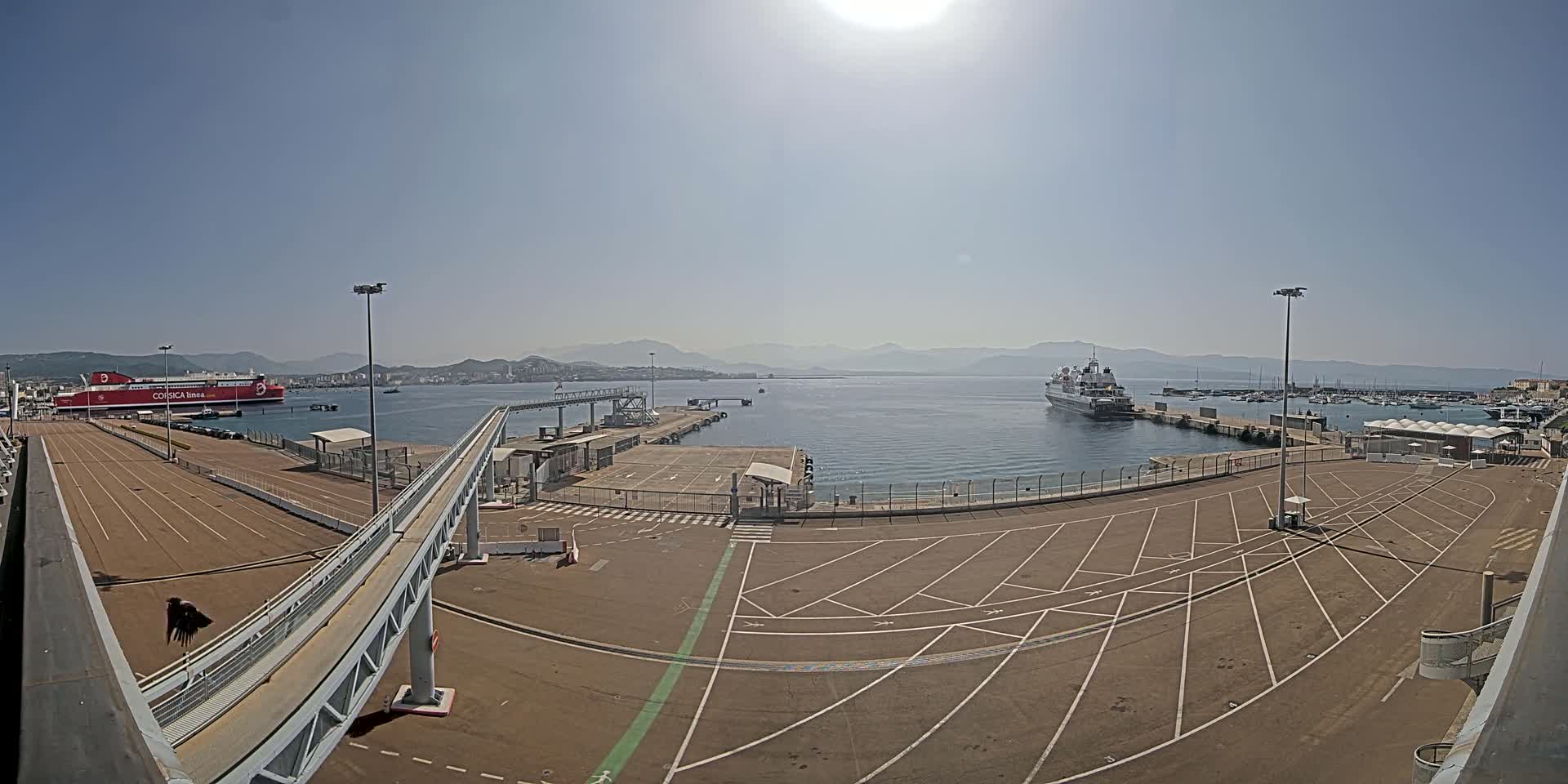 A sunny day at a port shows two large ships docked, one red and one white, with an empty parking lot and a walkway leading to a gangway.