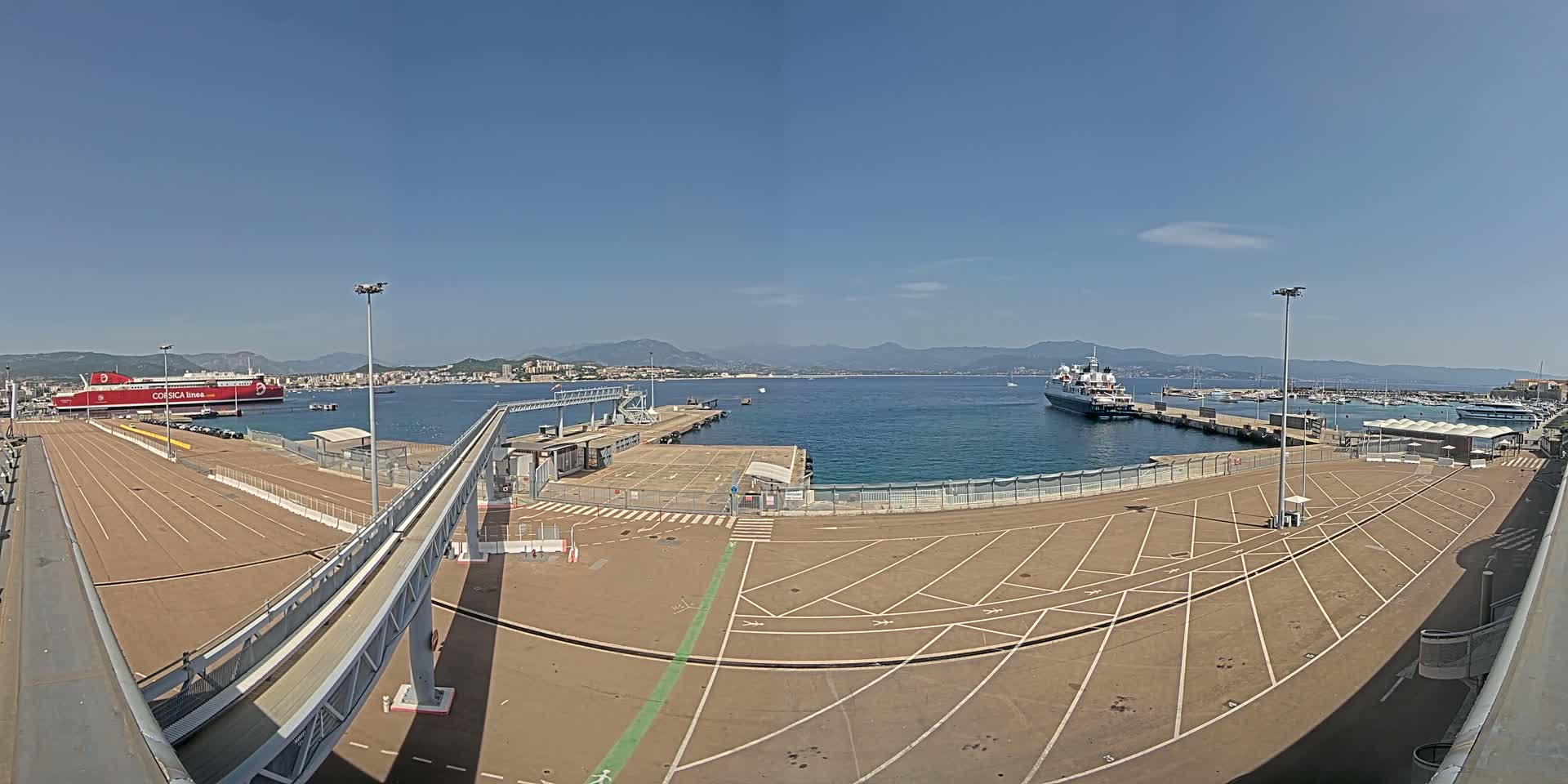 A mostly empty port area with a large red ferry docked, another ship in the water, and distant mountains under a clear blue sky.