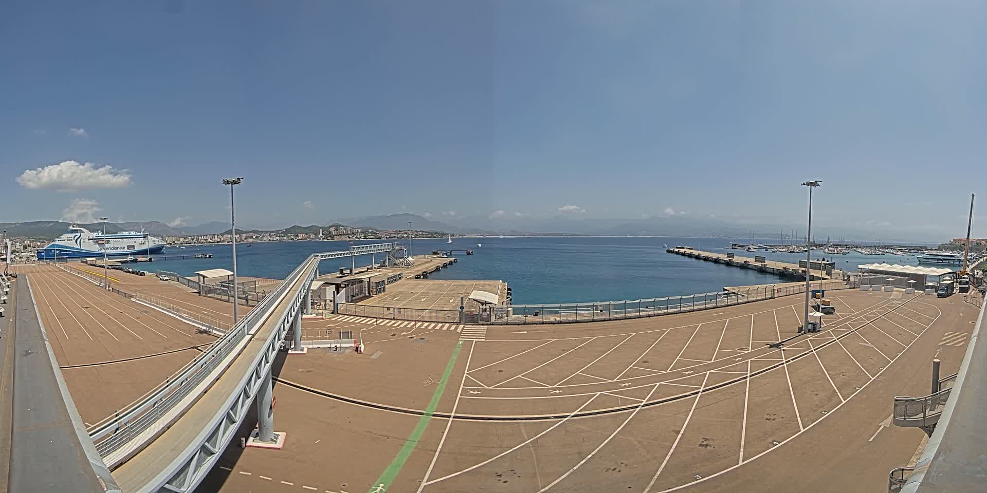 A ferry is docked at a port on a sunny day with clear skies and calm water, and mountains are visible in the distance.