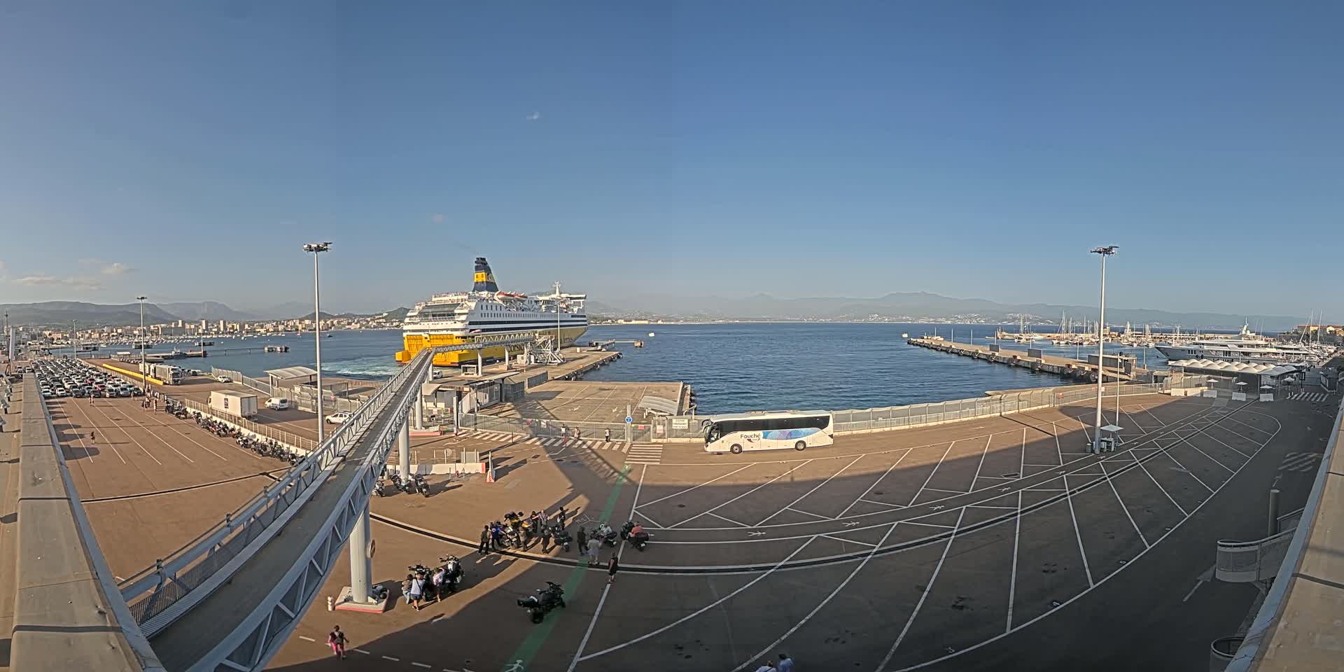 A large ferry is docked at a port on a sunny day, with numerous vehicles parked in the surrounding area and a distant mountain range visible.