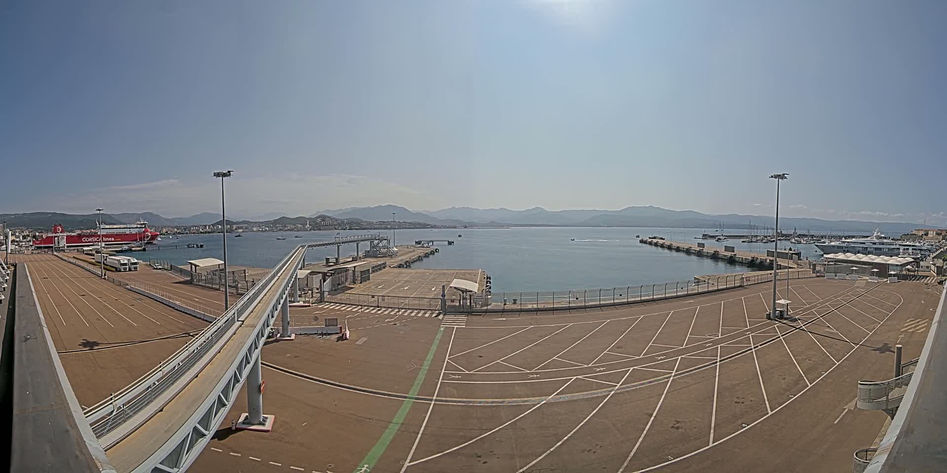 A large empty parking lot at a ferry terminal overlooks a calm harbor with docked boats and a ferry, all under a clear, sunny sky with distant mountains visible in the background.