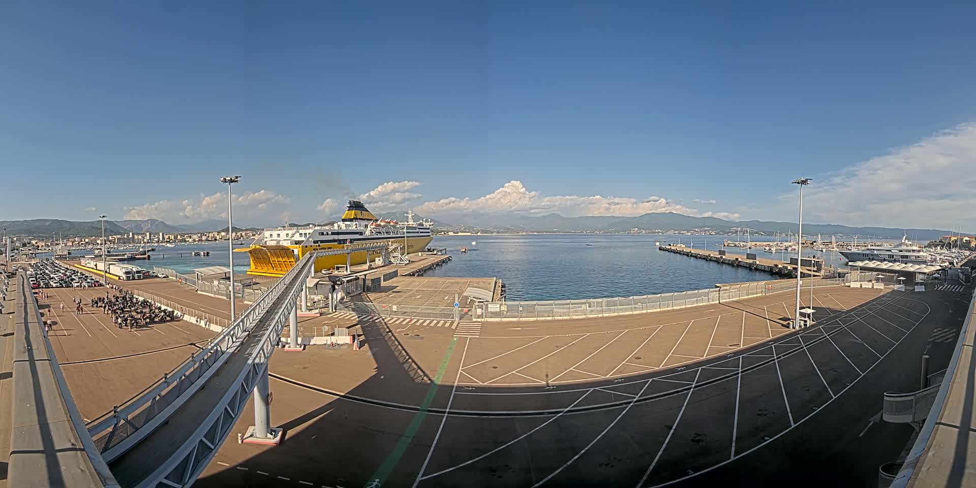 A large yellow ferry is docked at a port under a mostly clear sky with some scattered clouds and distant mountains visible in the background.