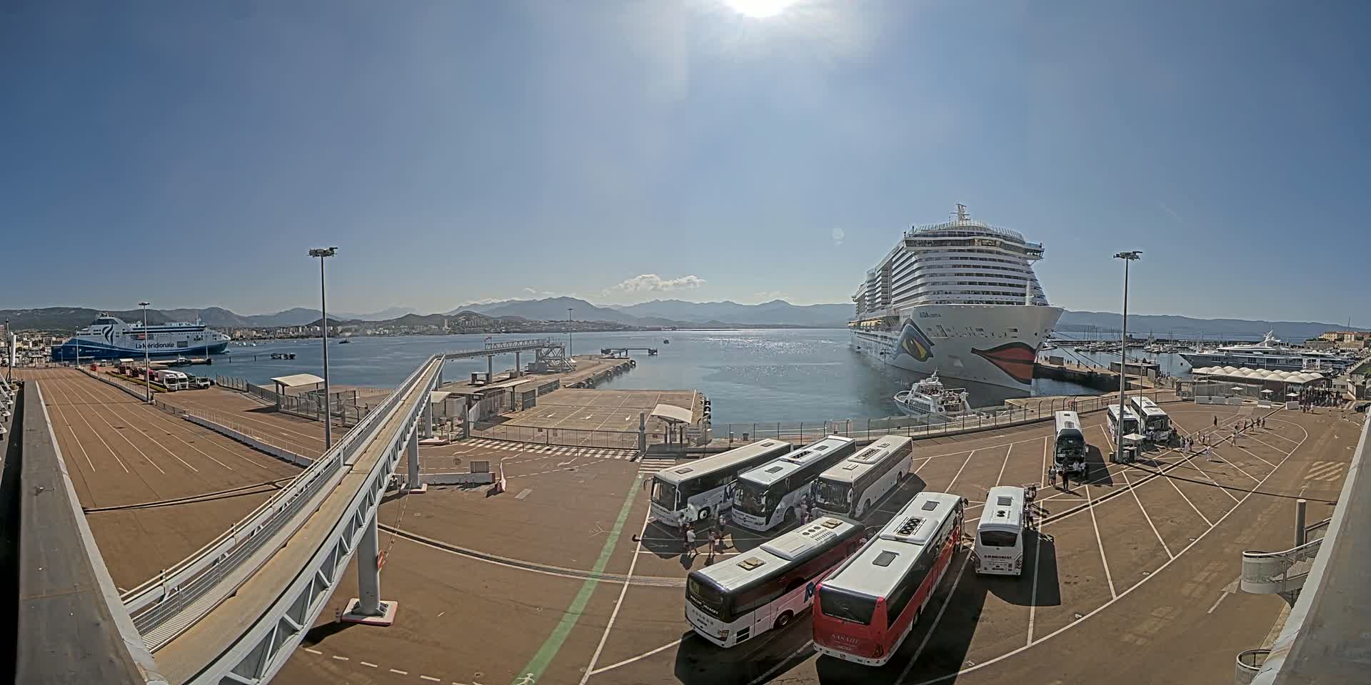 A cruise ship and a ferry are docked at a port on a sunny day, with several tour buses parked nearby.