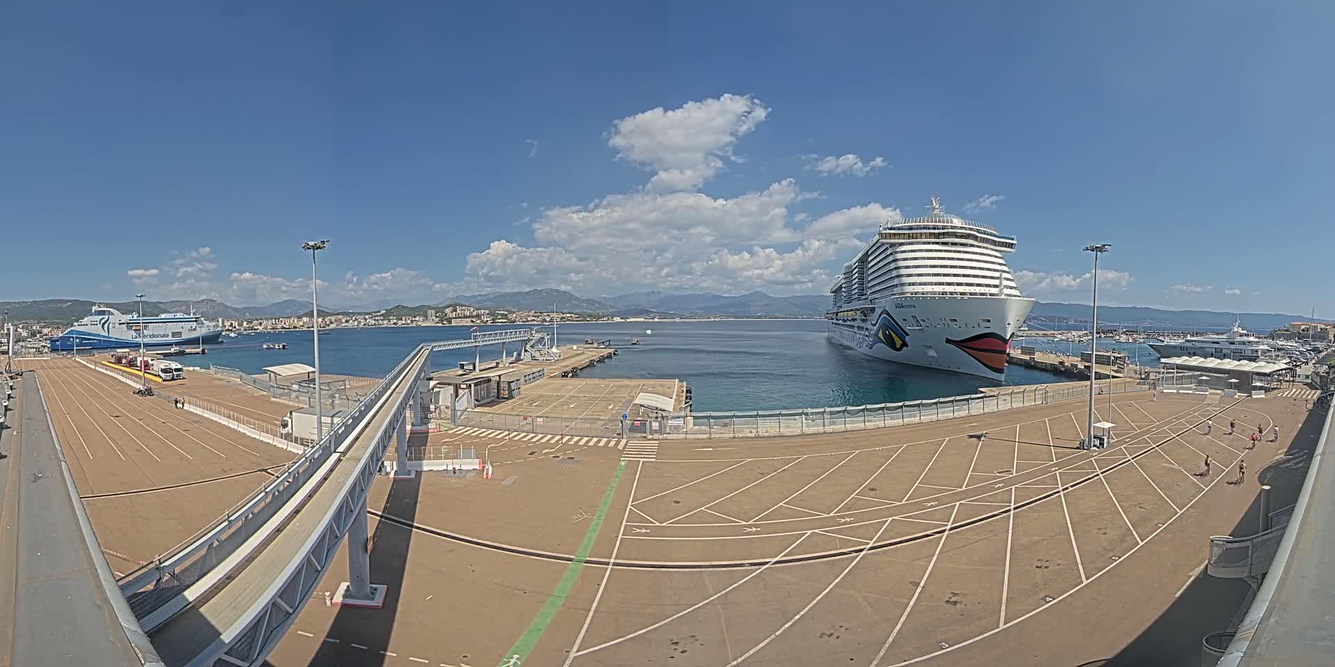 A cruise ship and ferry are docked at a port on a sunny day with partly cloudy skies and mountains in the background.