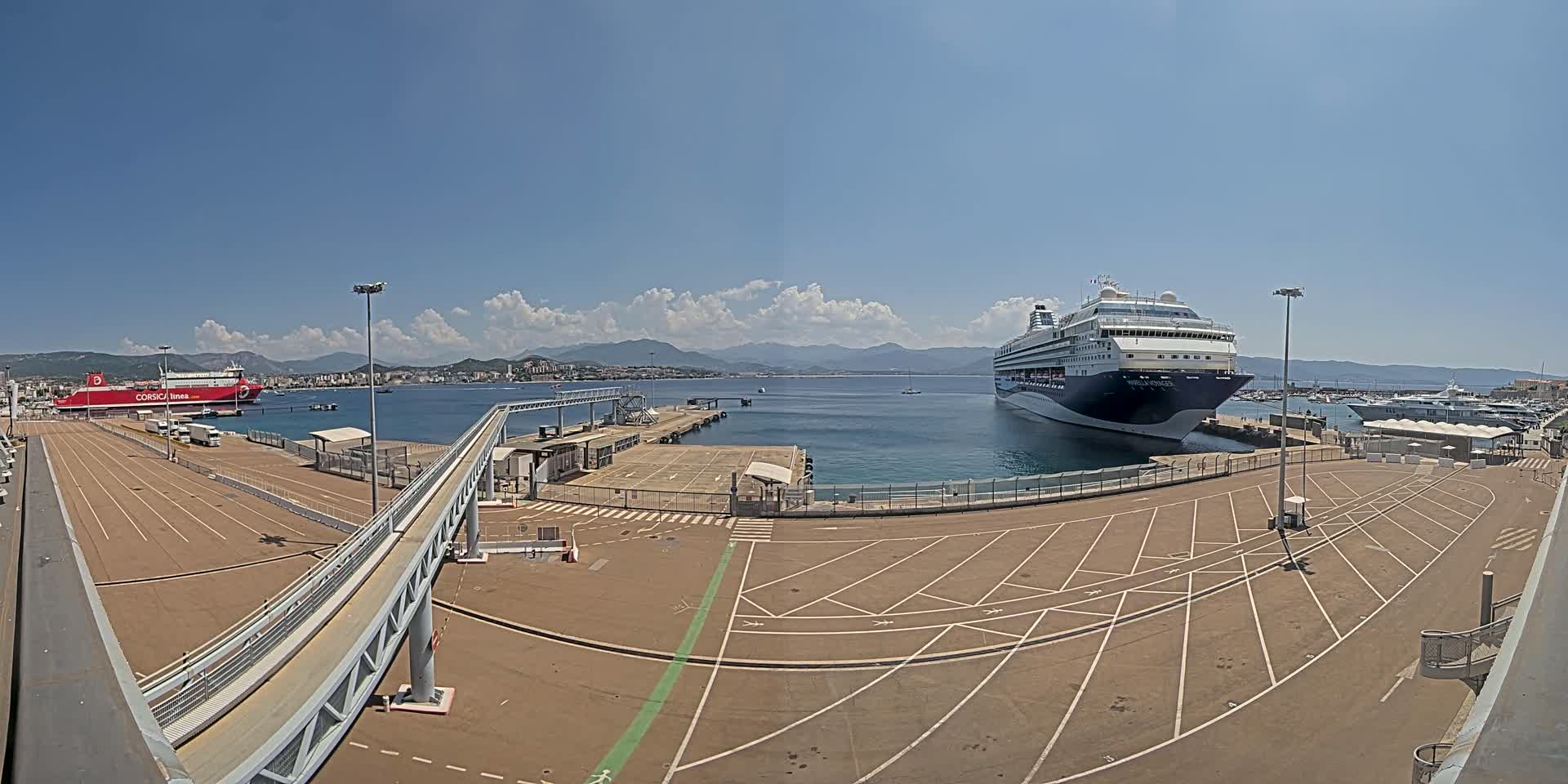 A cruise ship and a ferry are docked at a port on a sunny day with a partly cloudy sky and mountains in the background.