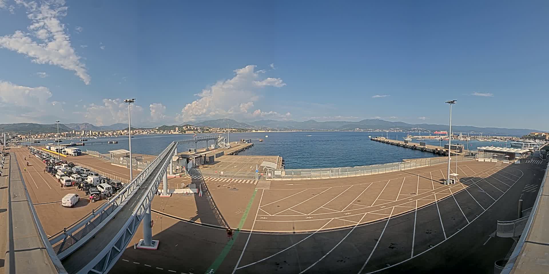 A mostly sunny day at a ferry terminal with a large empty parking lot, a walkway leading to a dock, and numerous vehicles parked near the terminal building; mountains and a body of water are visible in the background.