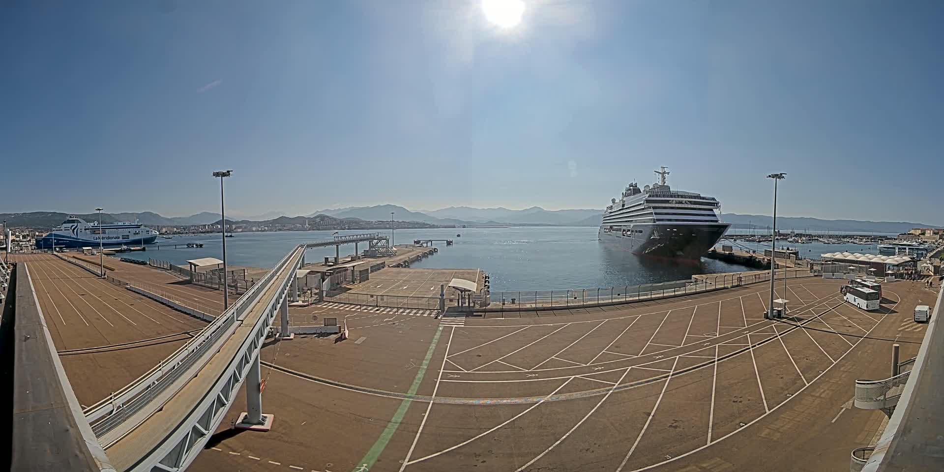 A large cruise ship and a ferry are docked at a port on a sunny day, with mountains visible in the distance.