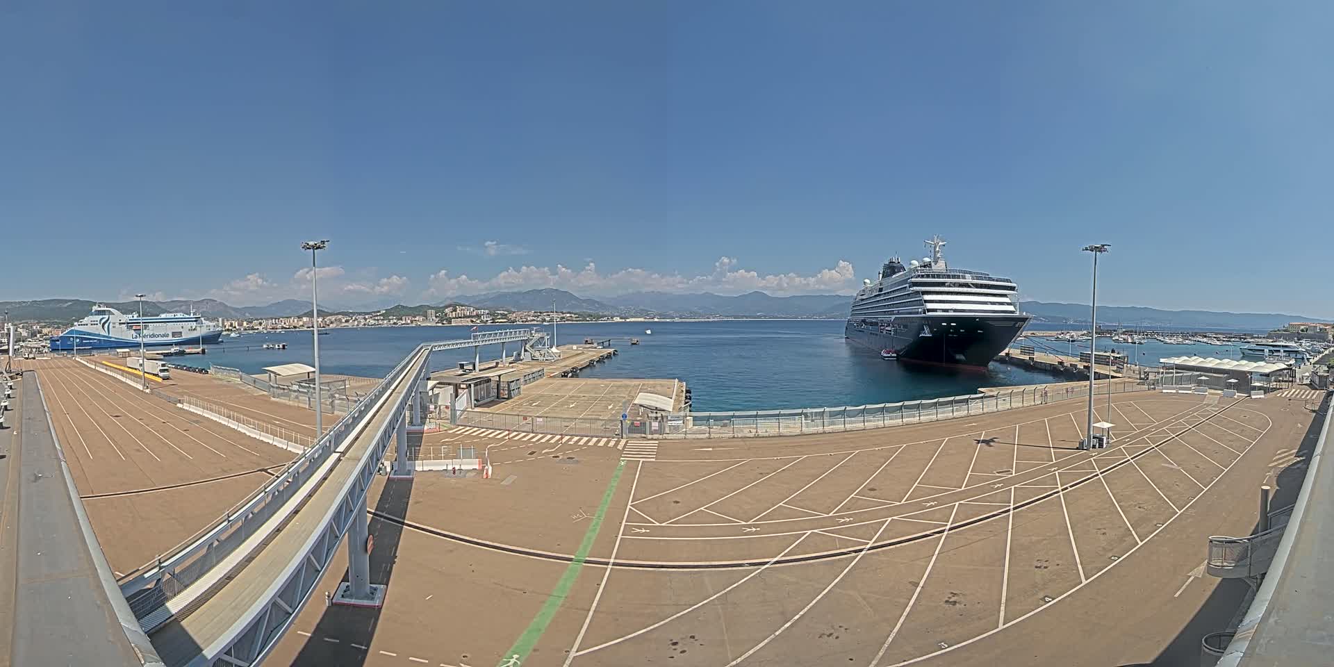 A large cruise ship and a ferry are docked at a port on a sunny day, with mountains visible in the distance.