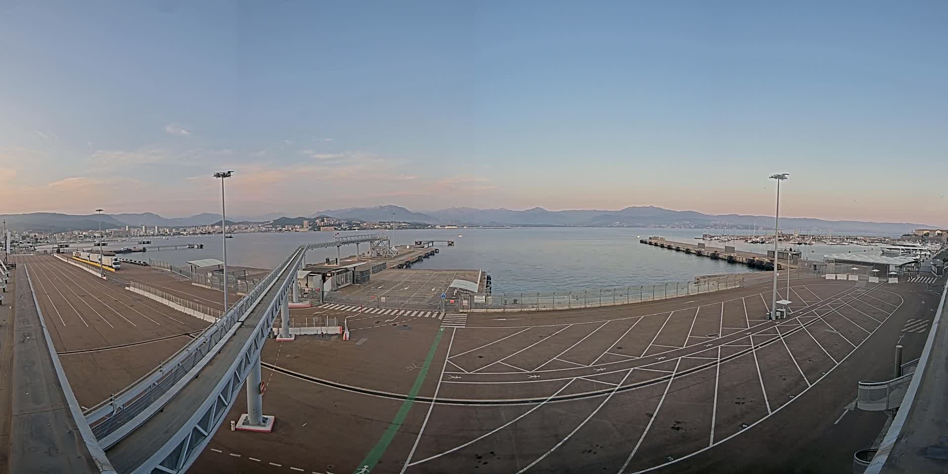 An empty port area with a walkway and parking lot overlooks a calm body of water and distant mountains under a clear, twilight sky.