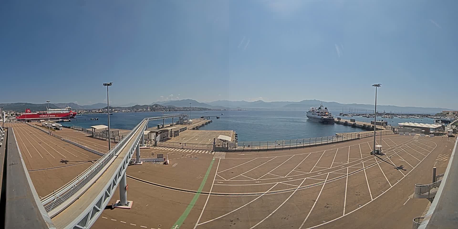 A sunny day at a port shows a large empty parking lot next to a dock with a passenger ferry and a cruise ship in the harbor, mountains in the background.