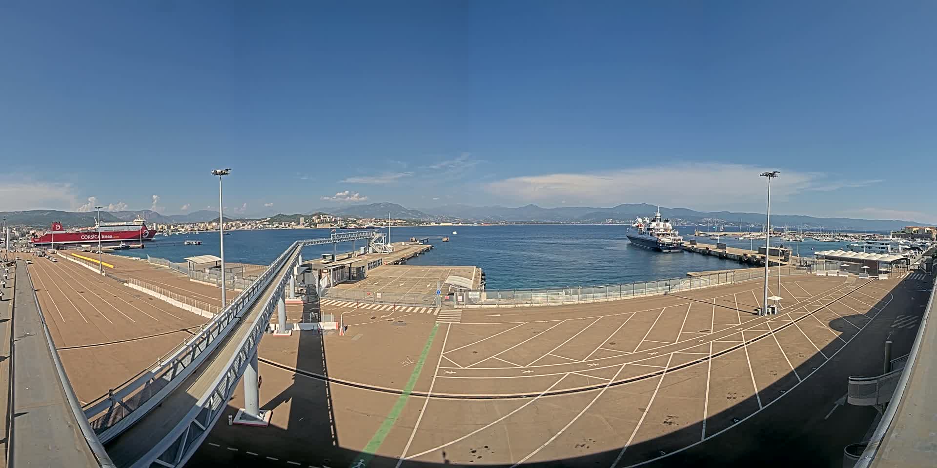 A large, empty parking lot at a port overlooks a calm body of water with docked ships and a distant mountainous coastline under a clear, sunny sky.