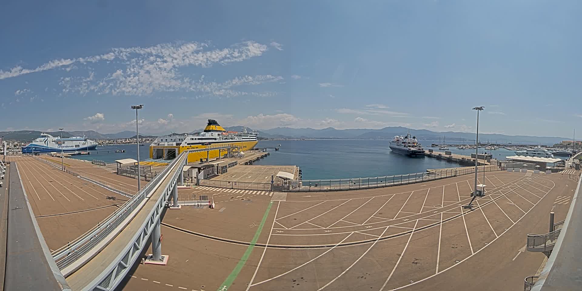 A large empty parking lot at a ferry terminal overlooks a calm harbor with several docked ferries and boats under a clear, sunny sky.