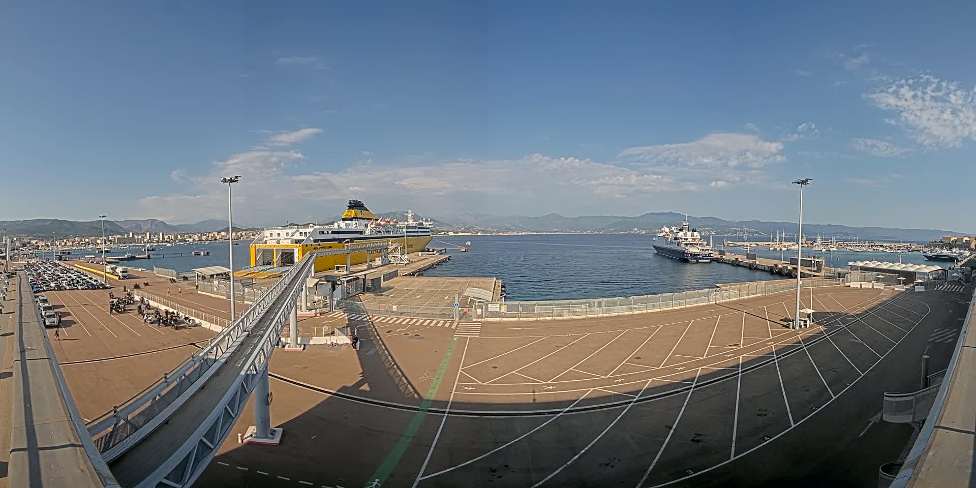 A large yellow ferry is docked at a port on a sunny day, with another ship visible in the distance and numerous cars parked in a nearby lot.