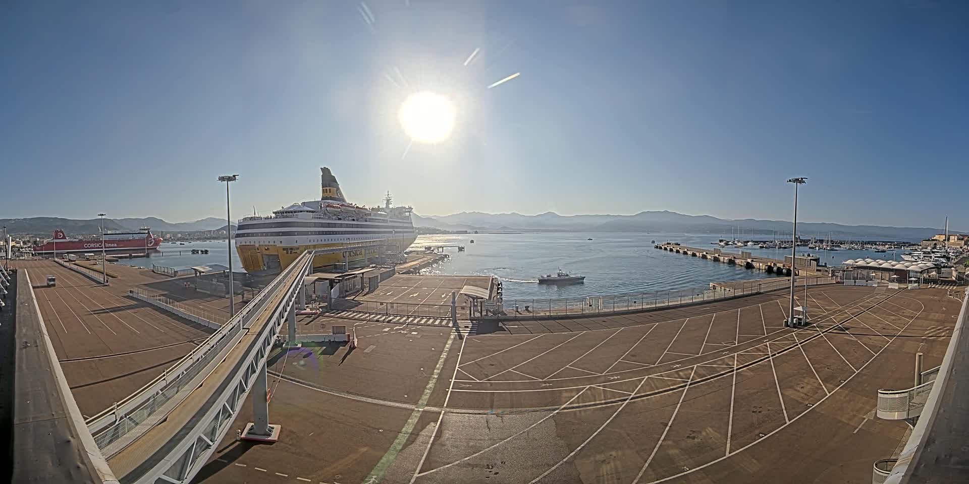 A large ferry is docked at a port on a sunny day with calm water, mountains in the distance, and numerous smaller boats moored nearby.