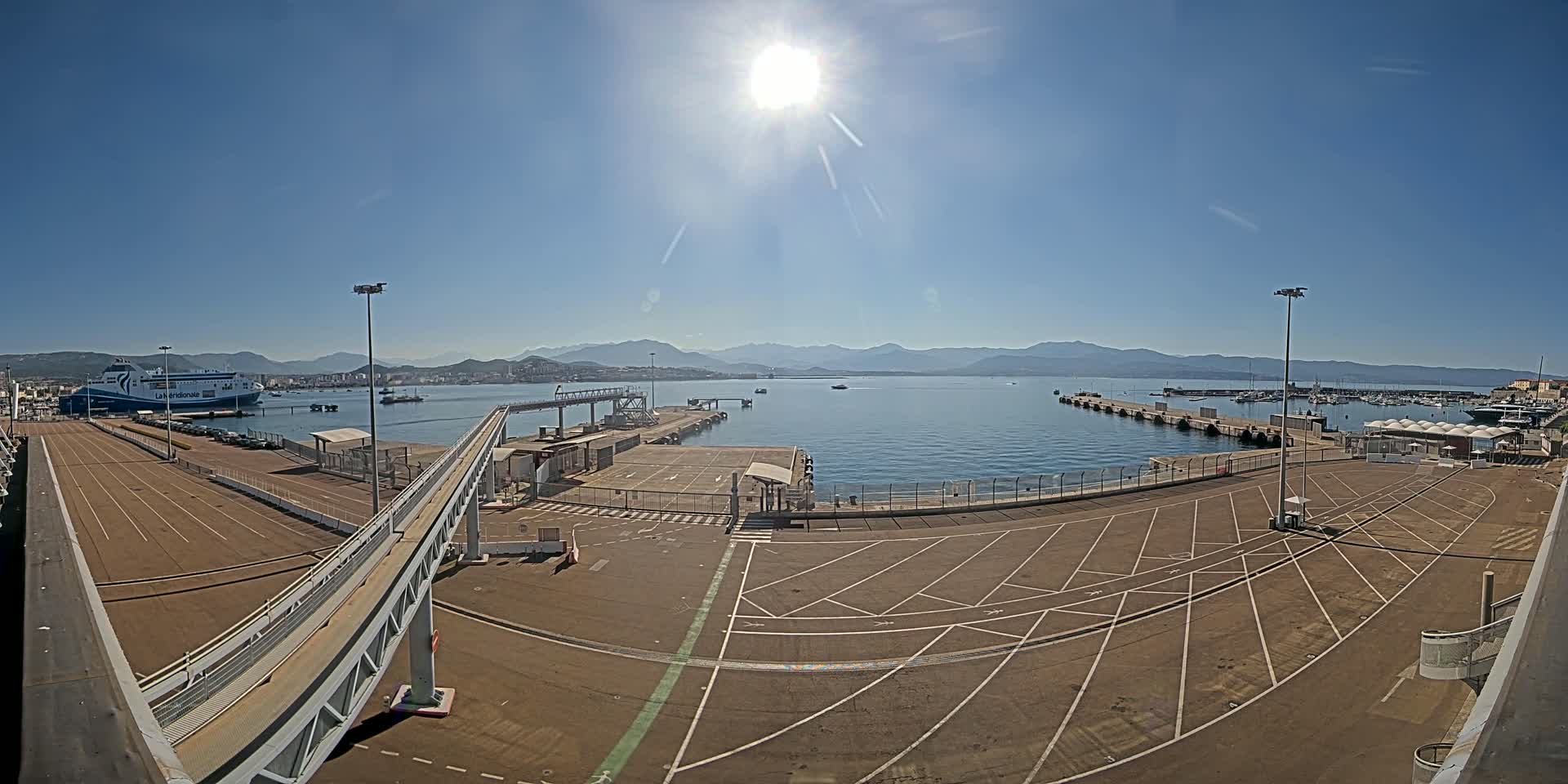 A sunny day at a ferry terminal shows a large ferry docked, a mostly empty parking lot, and a calm body of water with mountains in the distance.