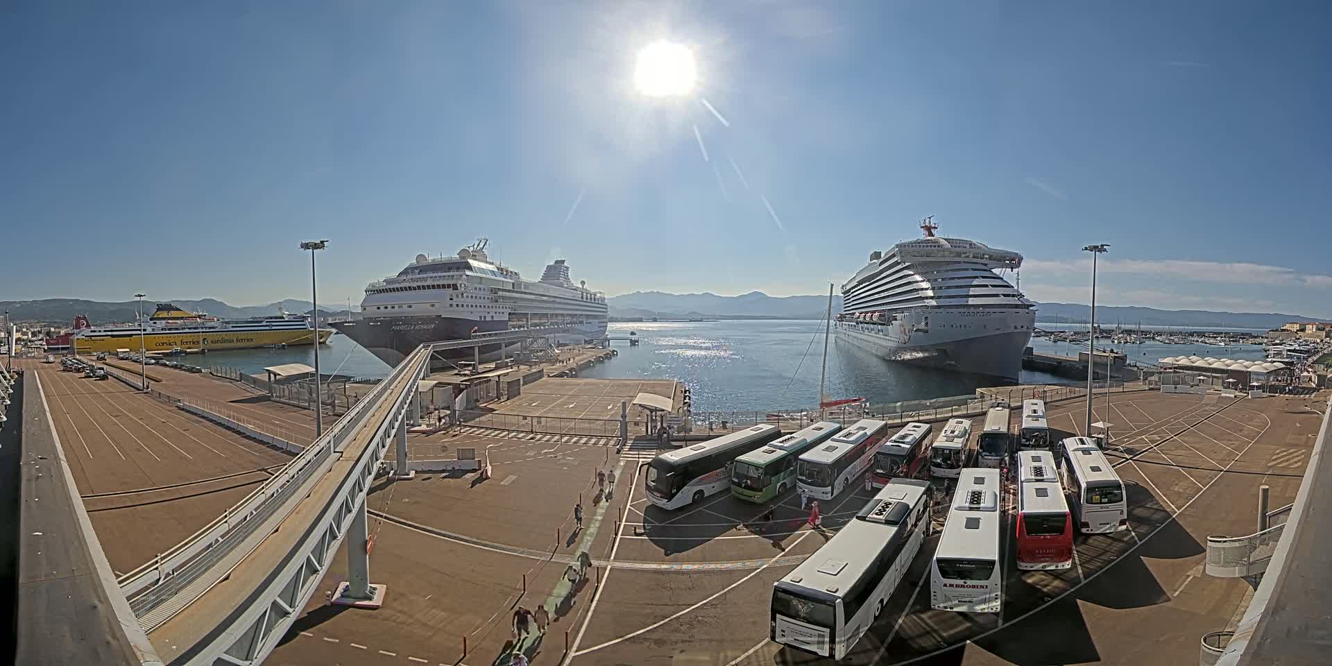Two large cruise ships and a ferry are docked at a port on a sunny day, with numerous tour buses parked nearby.