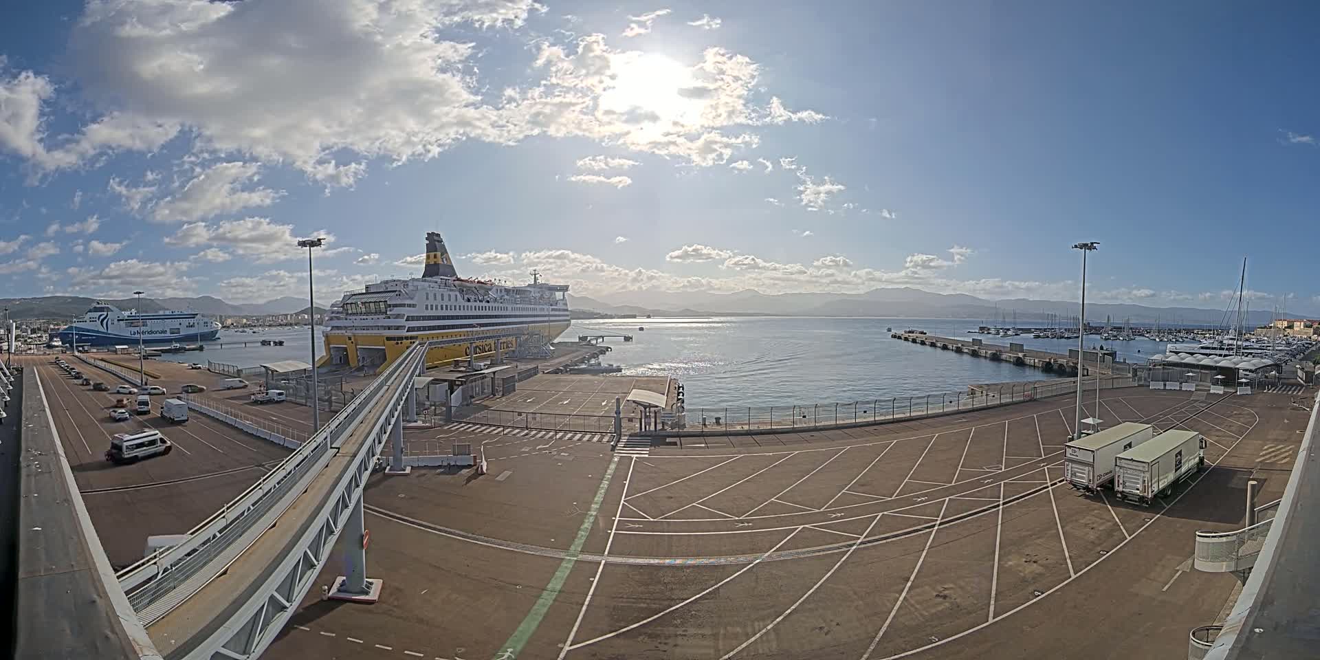 A large ferry is docked at a port on a sunny day with partly cloudy skies, with several vehicles parked nearby and a smaller ferry visible in the distance.
