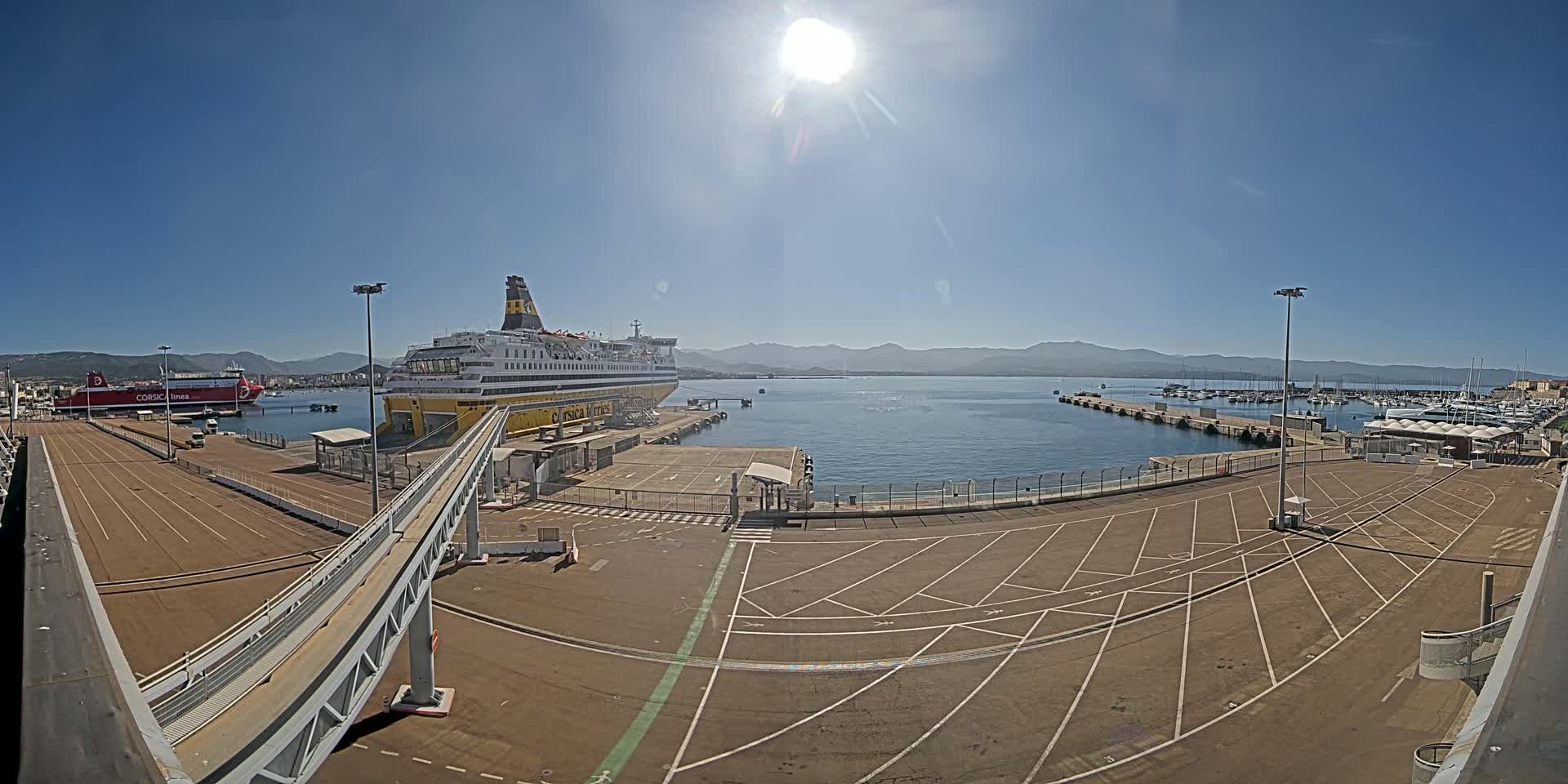 A large ferry is docked at a port on a sunny day, with another smaller vessel visible in the background and a large empty parking lot in the foreground.