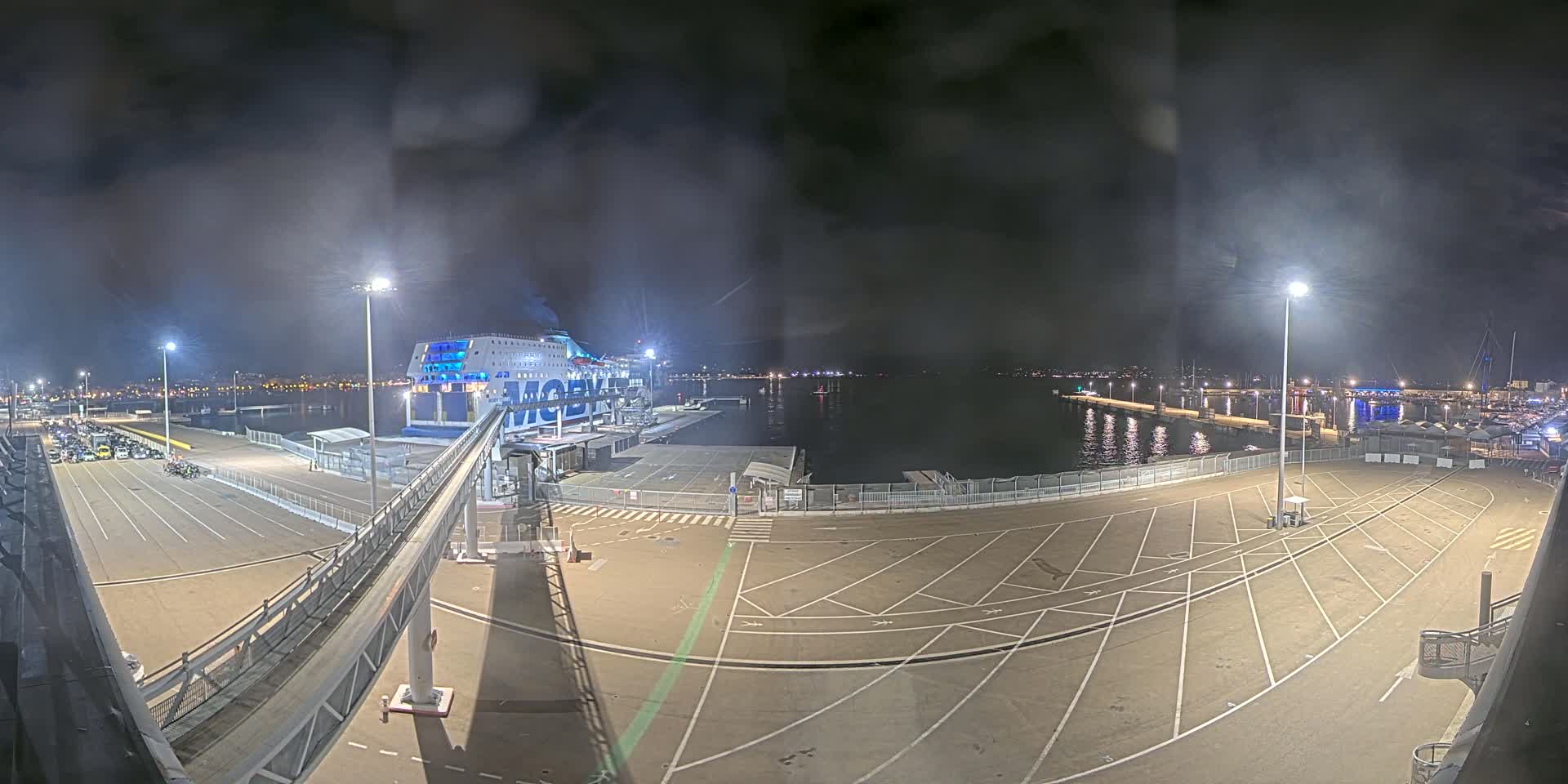 A large ferry is docked at a port at night under a hazy, dark sky, with a gangway connecting it to a mostly empty parking lot.