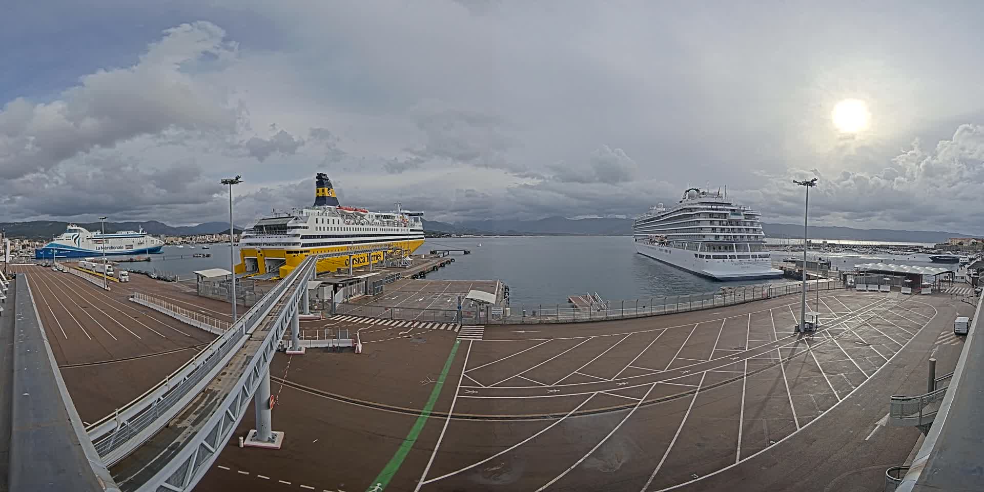 The image presents a harbor scene with a large yellow ferry, a prominent white cruise ship, and a distant blue ferry docked by concrete piers and access ramps, all illuminated by a bright sun breaking through an otherwise overcast sky.