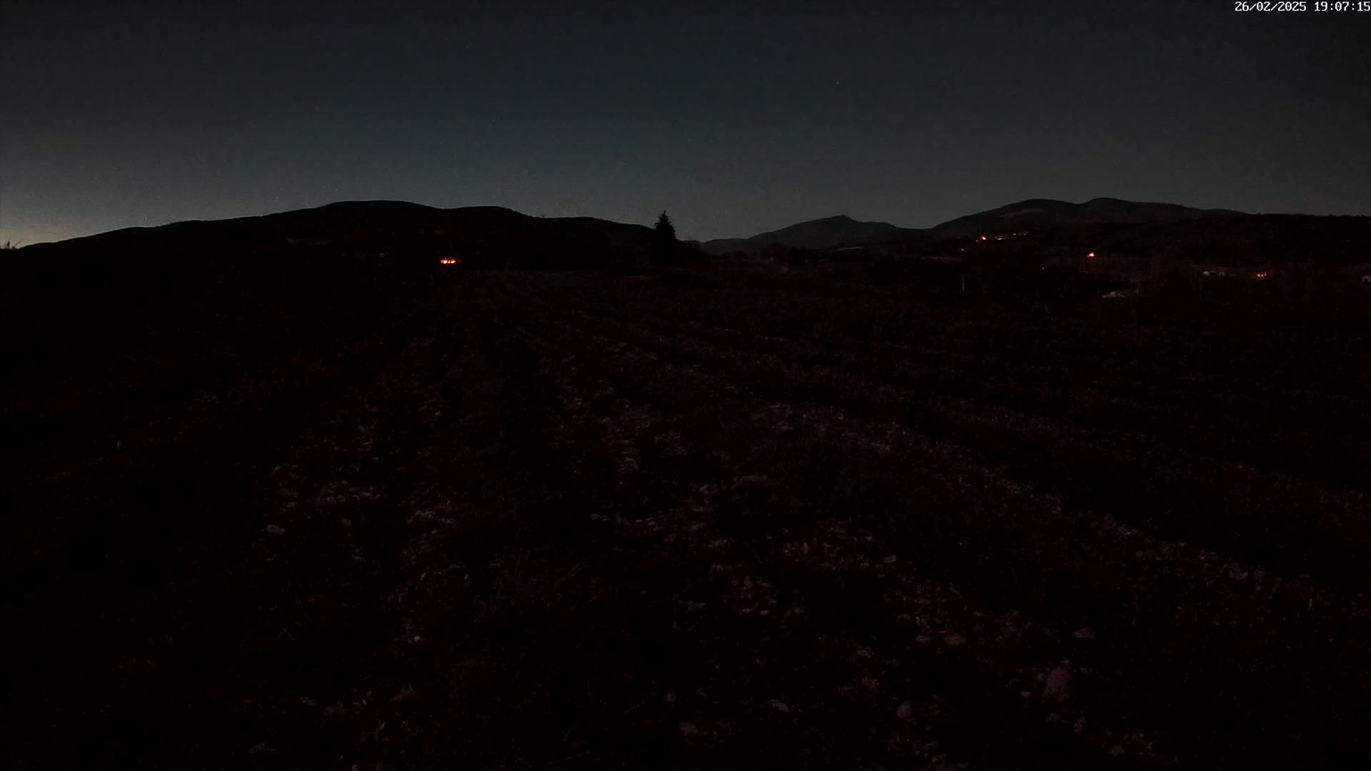 A dark, nighttime view of a field extending to distant, silhouetted mountains under a mostly clear sky.