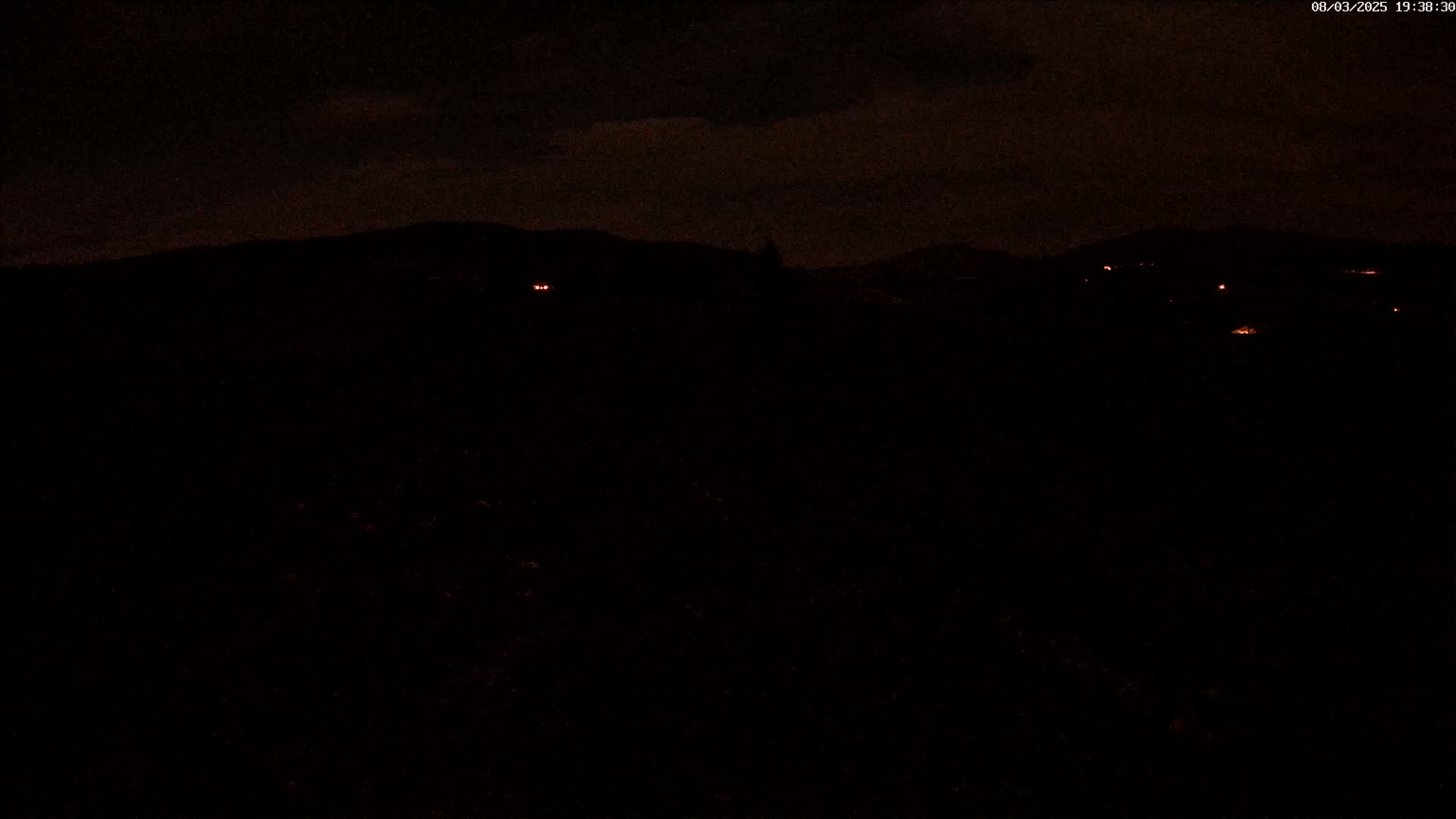 A nighttime view of dark hills and a few distant lights under a cloudy sky.
