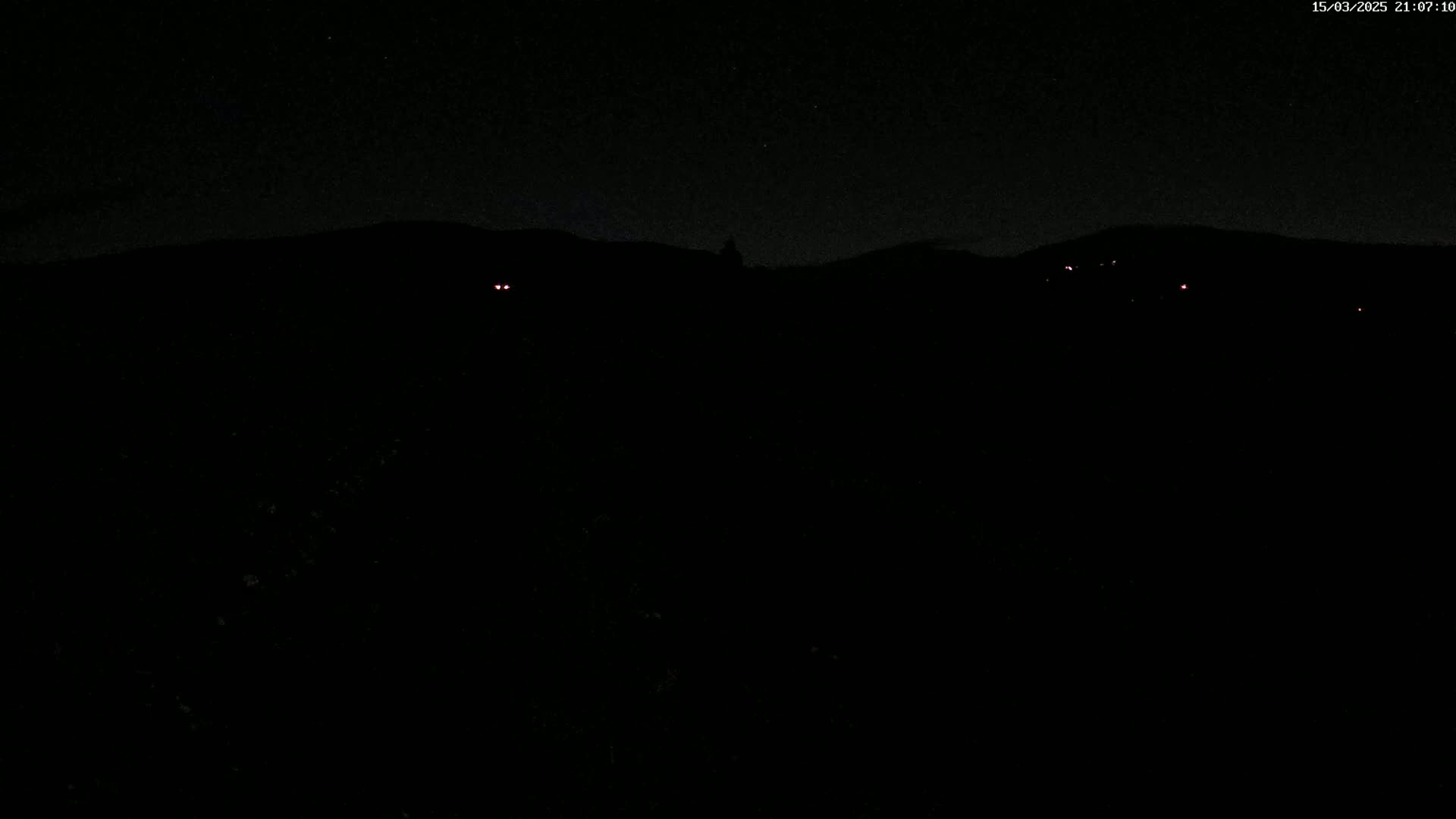 A dark, nighttime landscape is shown, featuring a distant, silhouetted hill line with a few faint red lights visible in the darkness.