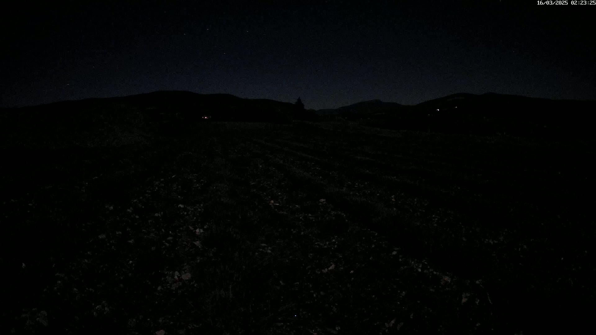 A dark, nighttime field stretches out before a silhouetted line of hills under a clear, starry sky.