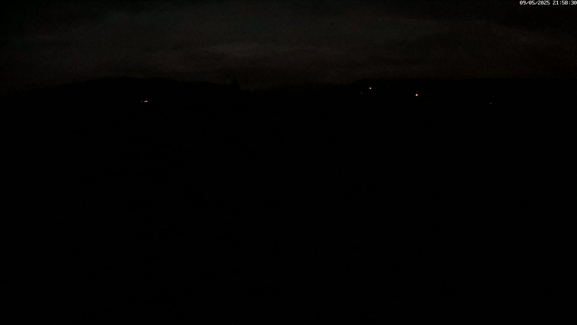 A dark, nighttime scene shows a distant, silhouetted mountain range with a few faint lights visible in the distance under a mostly cloudy sky.