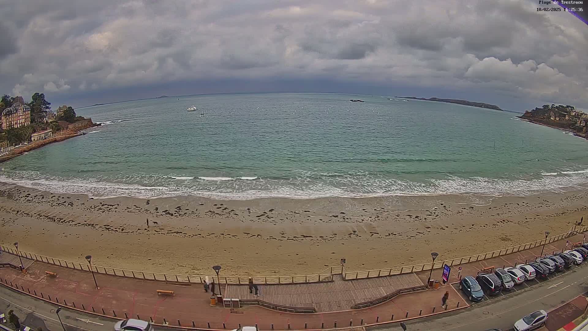 A sandy beach with gentle waves under a cloudy sky, bordered by a boardwalk, buildings, and a parking lot with cars.
