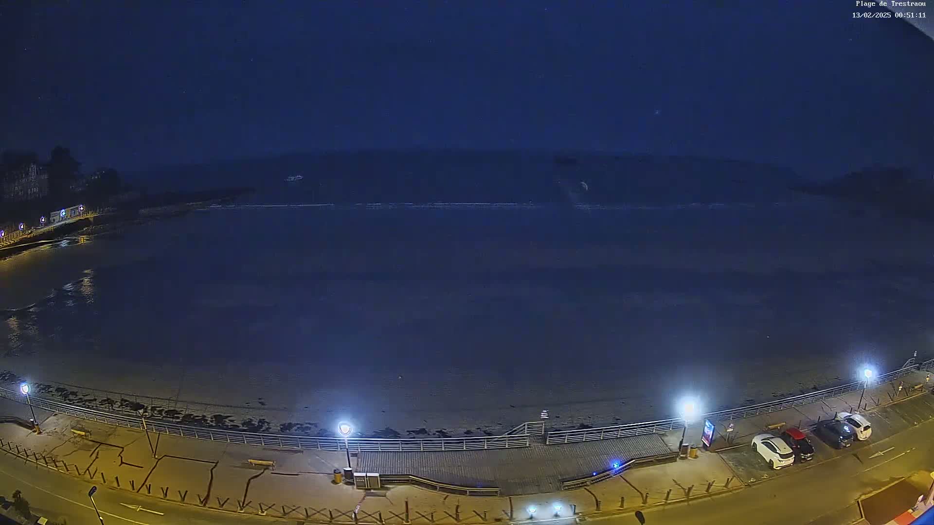 A dark, nighttime view of a mostly empty beach with a boardwalk, streetlights, and a few parked cars.