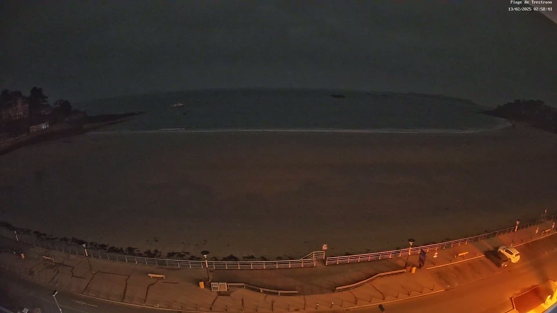 A dark, nighttime view of a mostly empty sandy beach bordered by a paved walkway with a few cars and people visible under dimly lit streetlights.