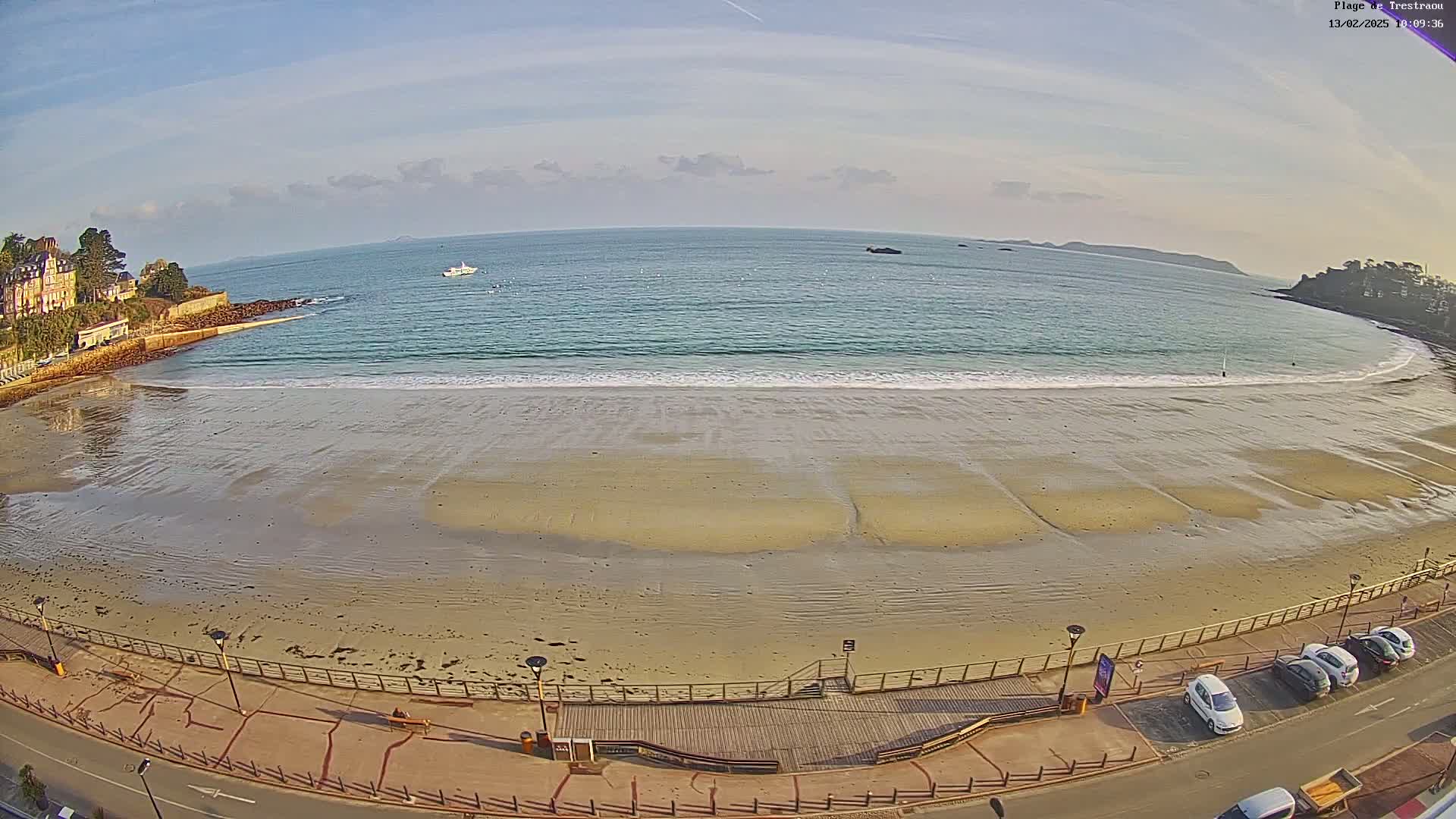 A sandy beach with calm ocean waves stretches along a shoreline, under a mostly clear sky with some clouds, and several cars parked nearby.