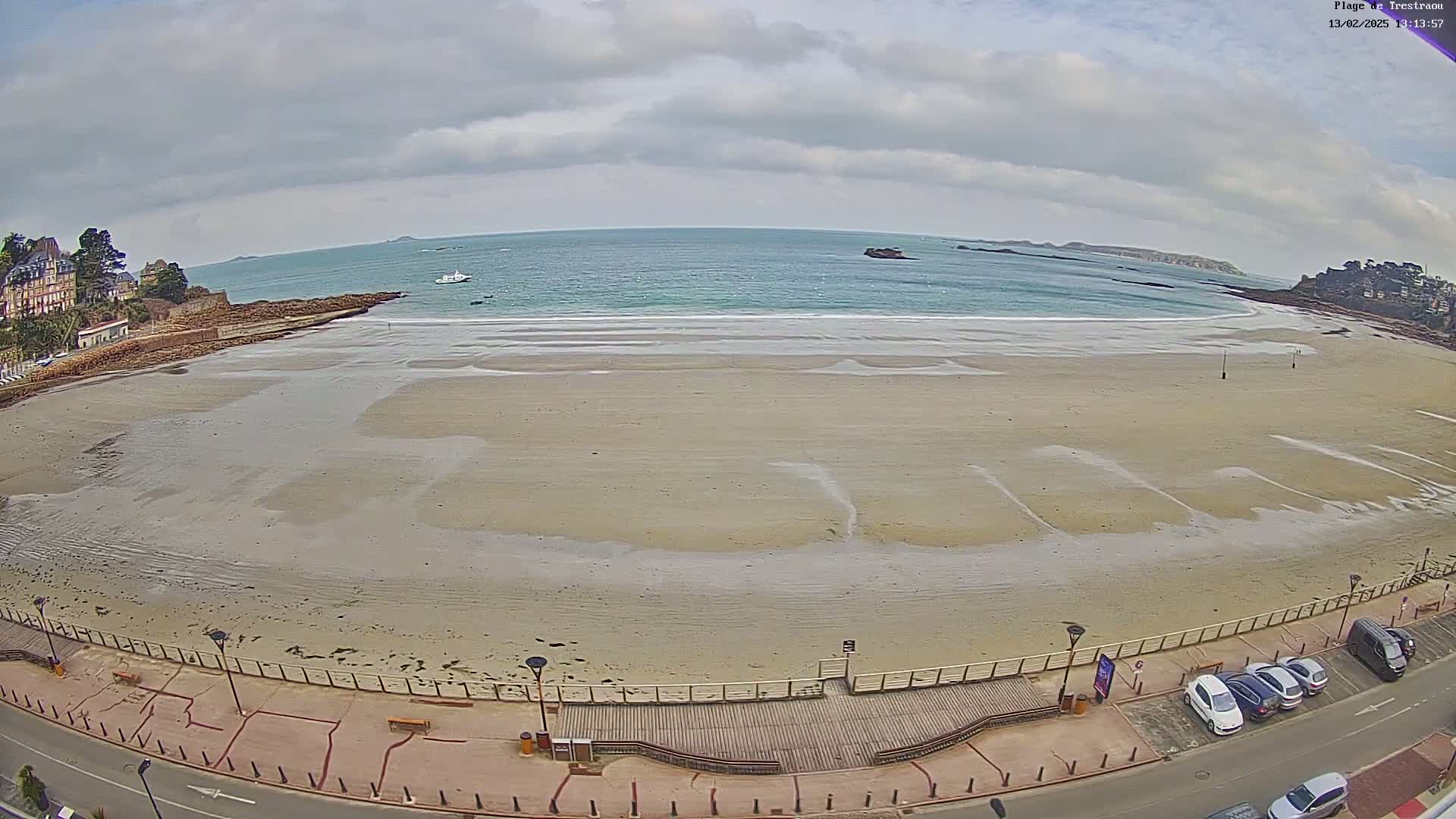 A wide sandy beach stretches towards a calm ocean under a cloudy sky, with a boardwalk and parked cars visible along the shore.