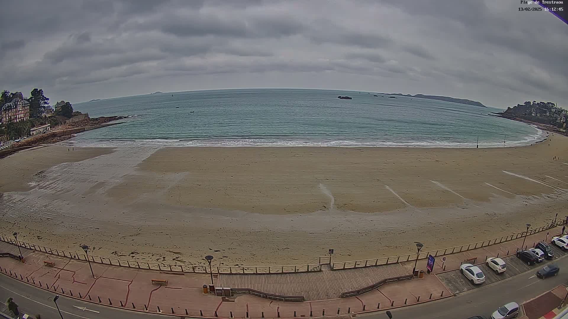 A sandy beach stretches along a calm, teal ocean under a cloudy sky, with a boardwalk, parked cars, and buildings visible along the shore.