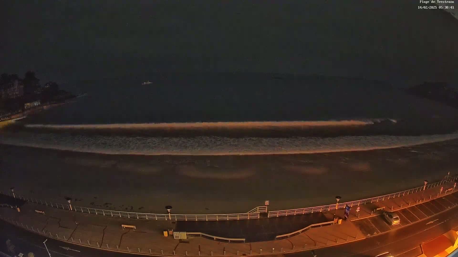 A long, gently rolling wave washes ashore at night on a dark beach, next to a dimly lit boardwalk and parking lot.