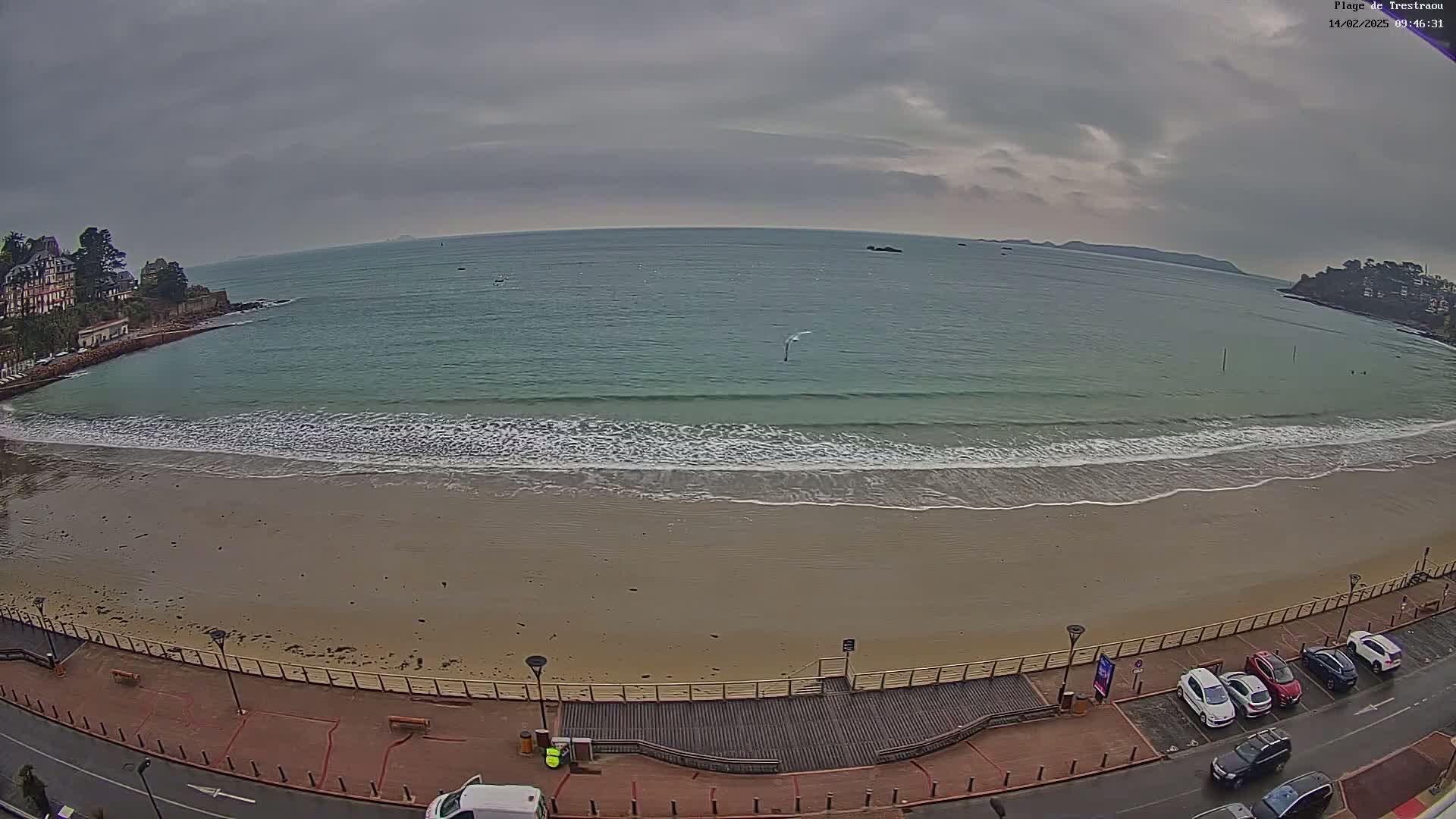 A sandy beach with gentle waves laps the shore under an overcast sky, bordered by a boardwalk and a street lined with parked cars.