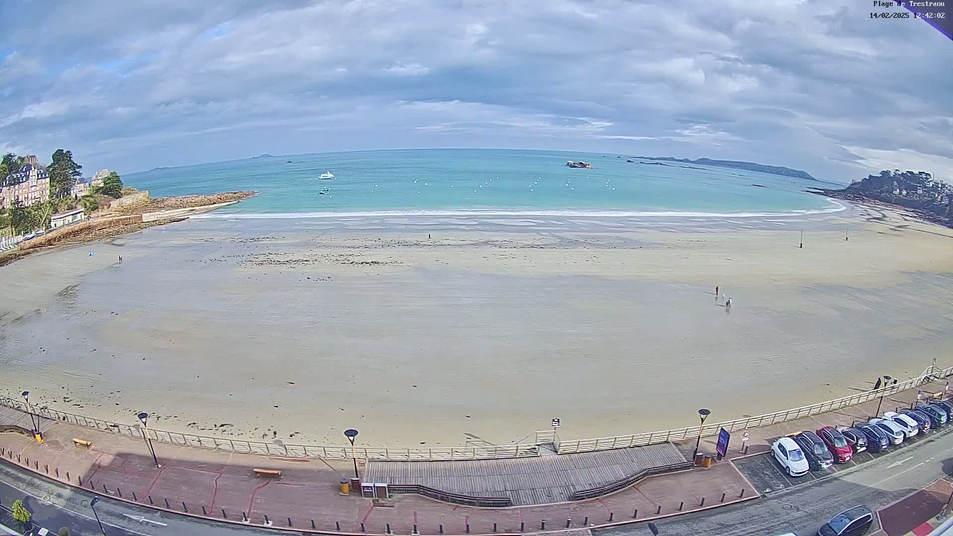 A mostly empty sandy beach stretches out to a calm, light teal ocean under a cloudy sky, with a boardwalk, parked cars, and buildings visible in the foreground.
