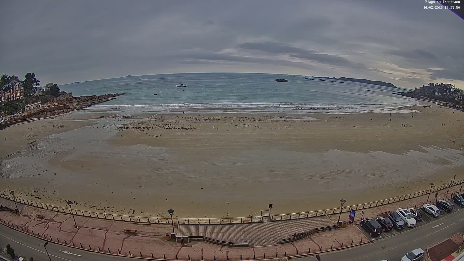 A wide sandy beach stretches along a calm ocean under an overcast sky, with a boardwalk and parked cars visible along the shore.