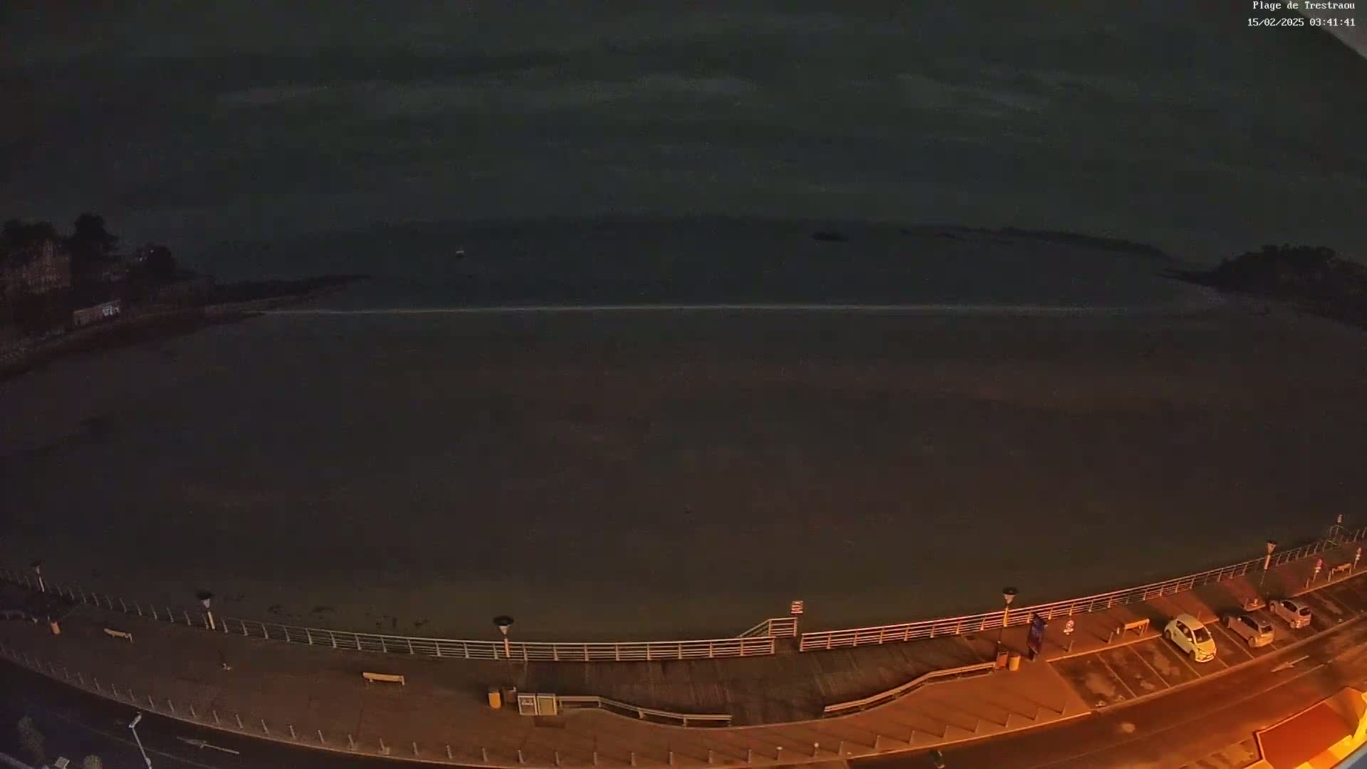 A dark, nighttime view of a mostly empty beach with a boardwalk and a few parked cars, under a cloudy sky.