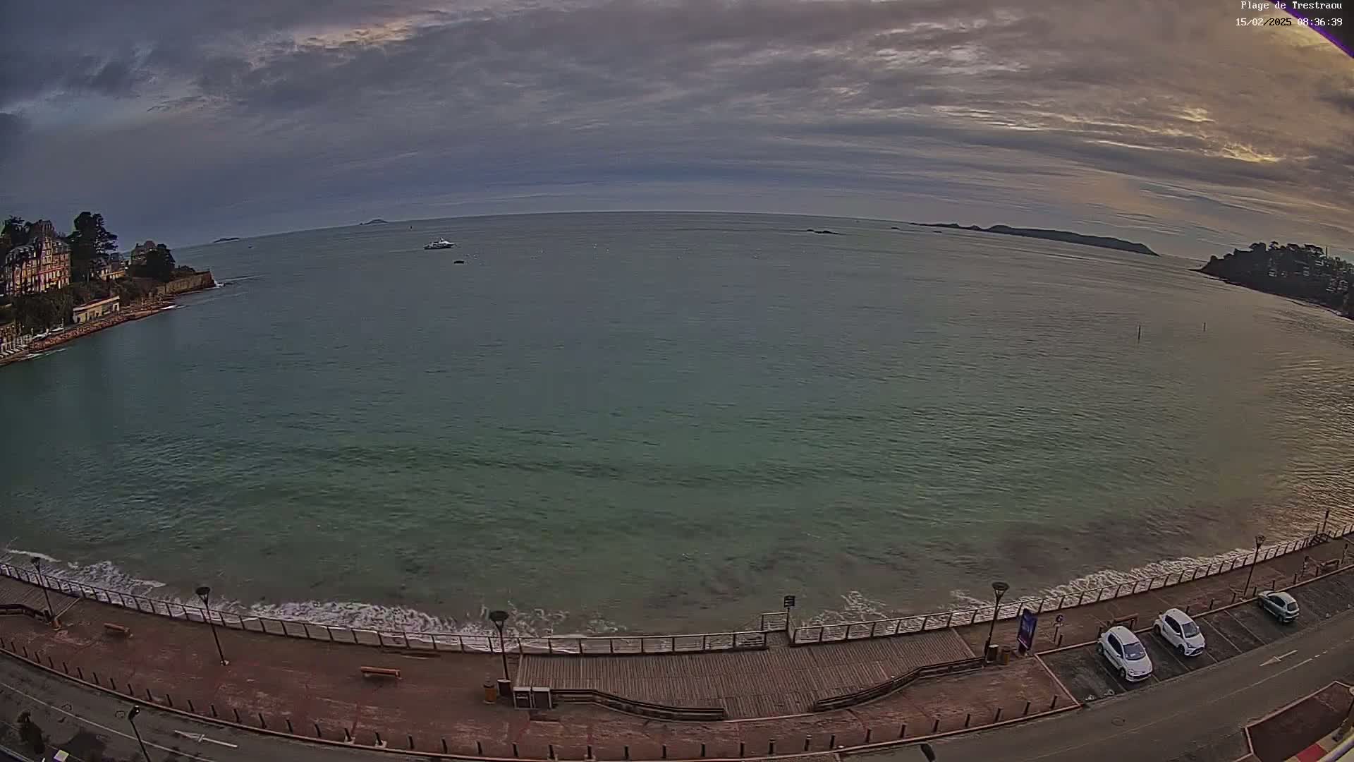 A wide shot of a calm, greenish-blue ocean with small waves lapping a boardwalk, overlooking a shoreline with buildings and several parked cars under an overcast sky.