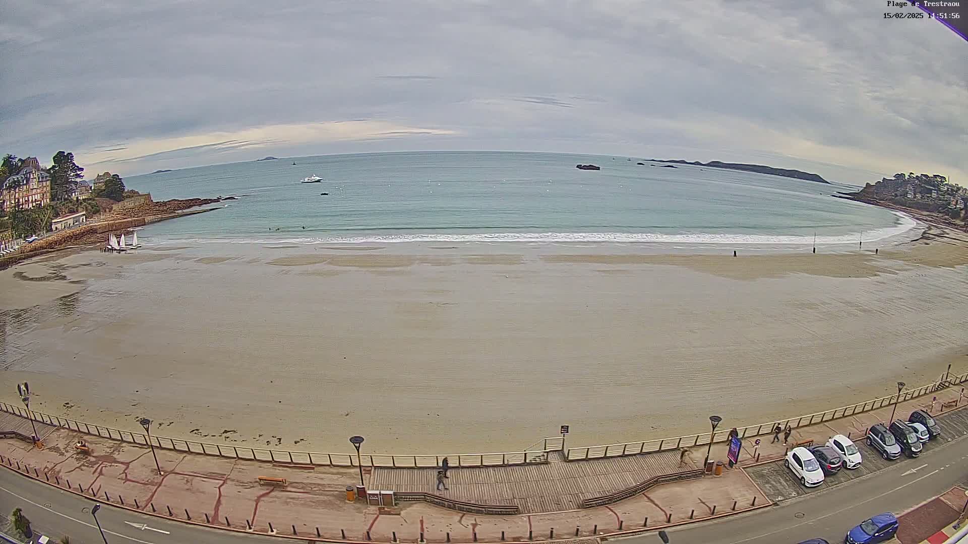 An overcast day reveals a wide sandy beach with a boardwalk and parked cars, all overlooking a calm ocean with small waves and distant landmasses.