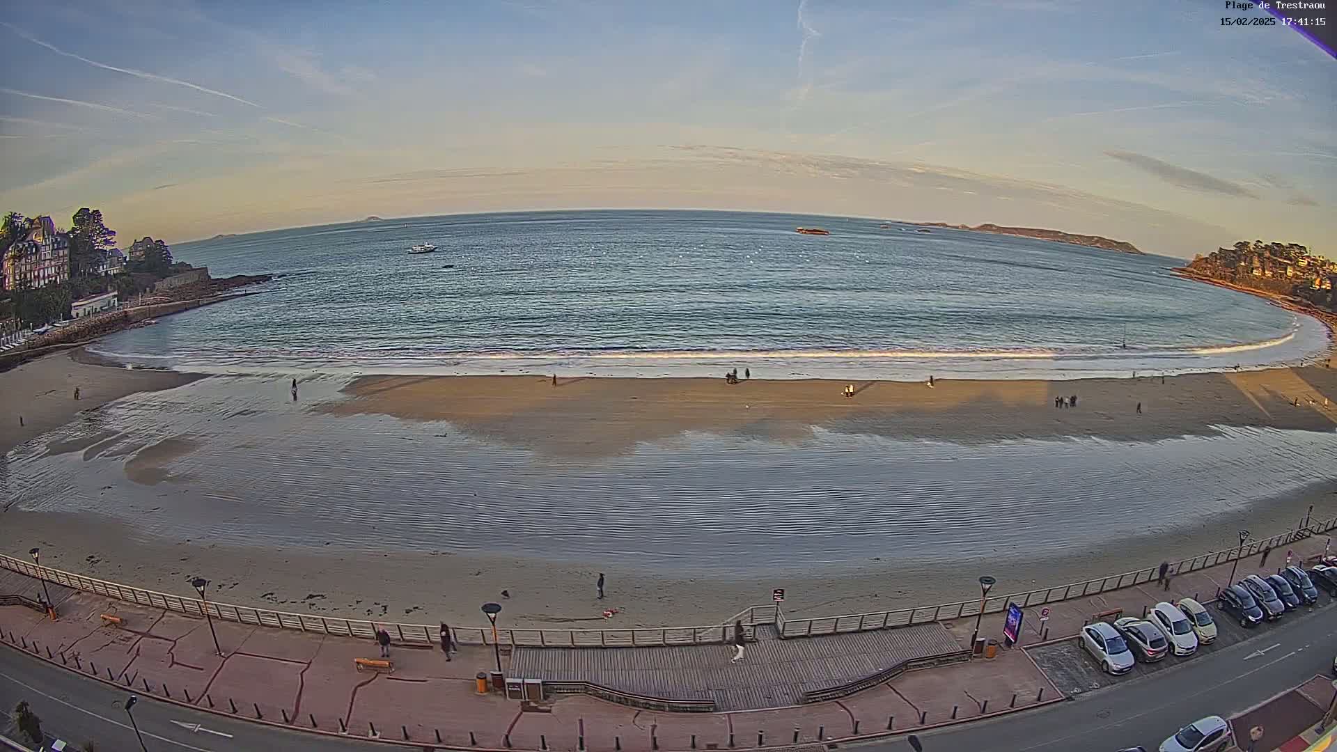 A sandy beach with a calm ocean under a clear, twilight sky, showing several people walking along the shore and cars parked along the road next to a boardwalk.