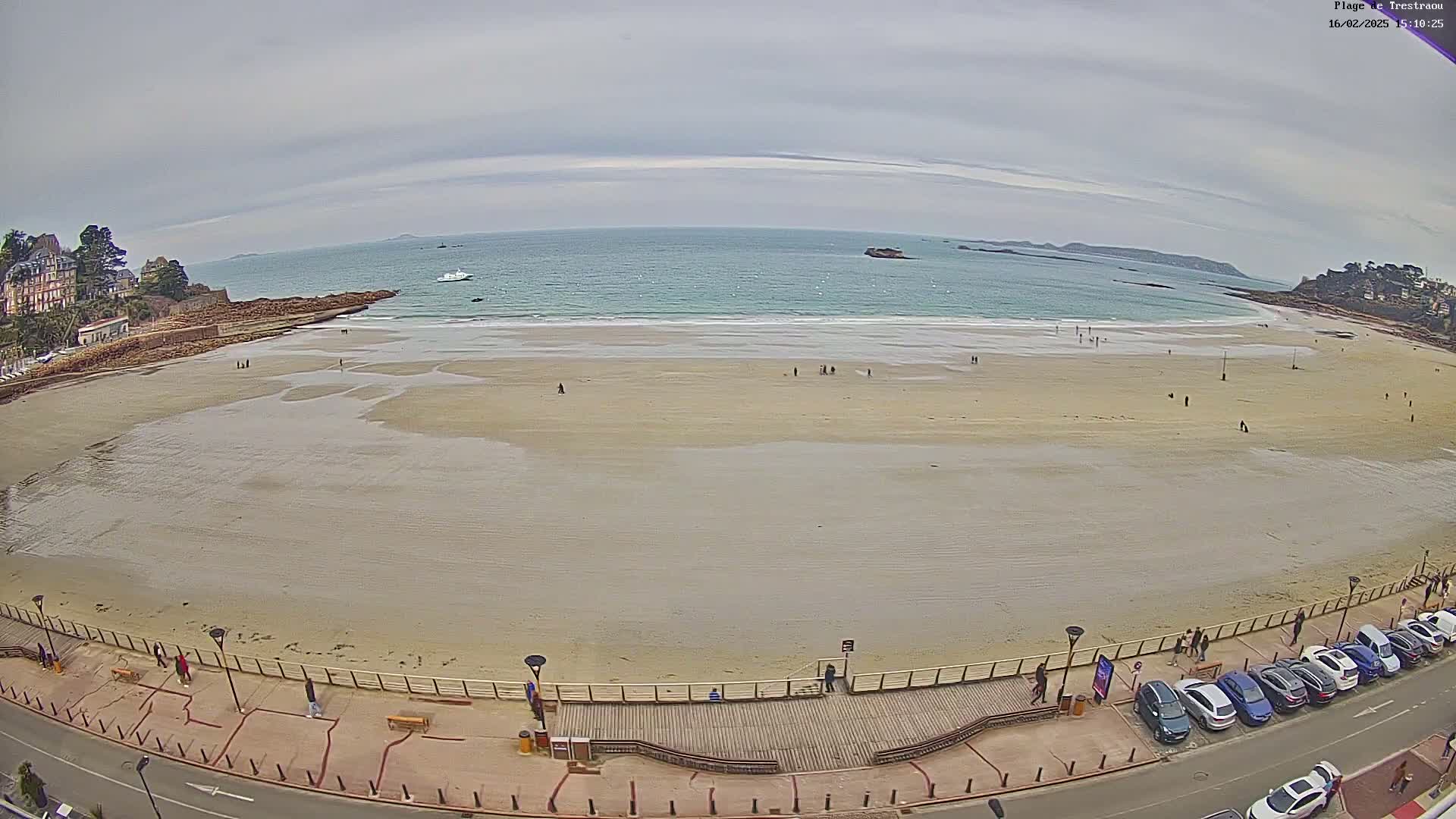 A wide shot of a sandy beach with a few people scattered across it, under an overcast sky, with calm water and several boats in the distance, and cars parked along a boardwalk next to the beach.