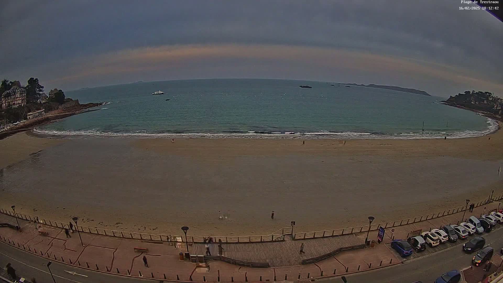 A wide sandy beach curves along a calm ocean under an overcast sky, with a few people scattered on the sand and cars parked along the roadway bordering the beach.