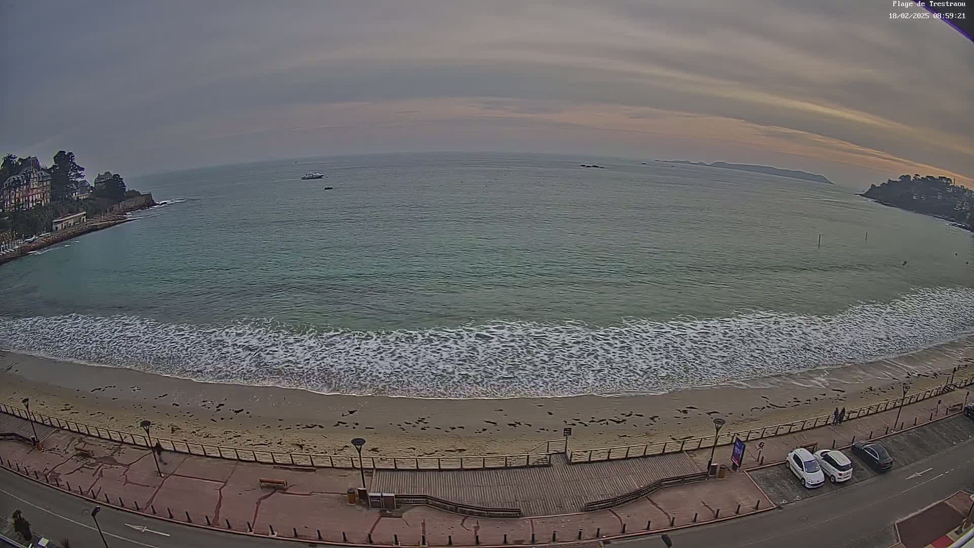 A sandy beach with gentle waves laps the shore under an overcast sky, with a road and parked cars visible along the beachfront.