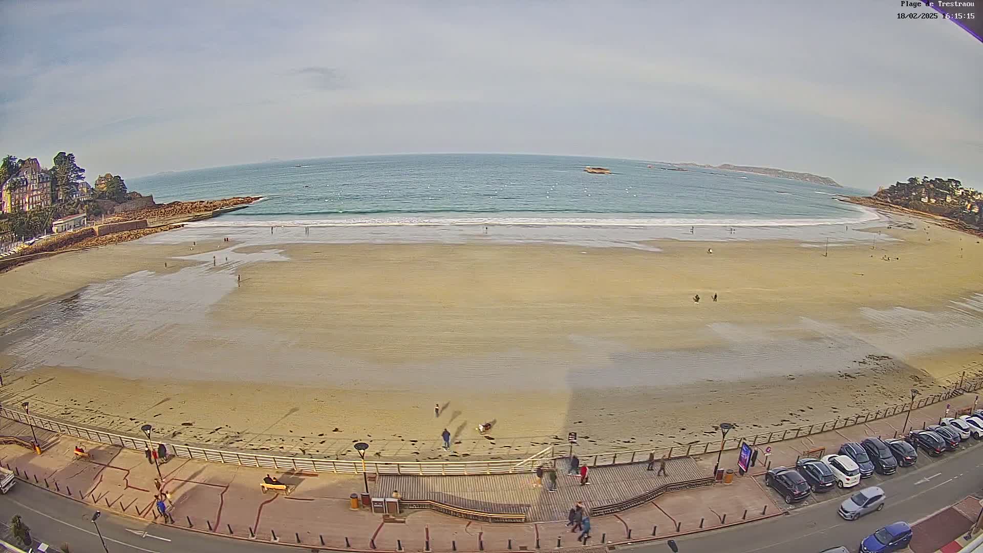 A wide sandy beach with a few people on it and a calm ocean with small waves under a mostly clear sky is viewed from above.