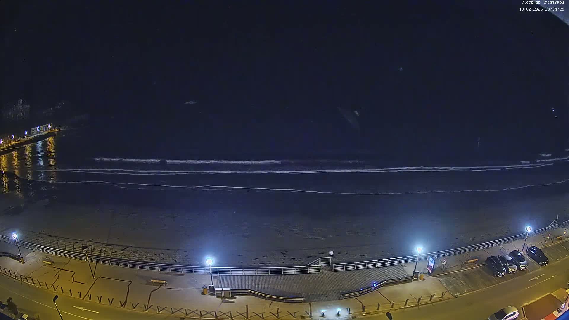 A nighttime, high-angle view shows a dark beach with small waves lapping the shore, a boardwalk with lights, and a few parked cars along a road.