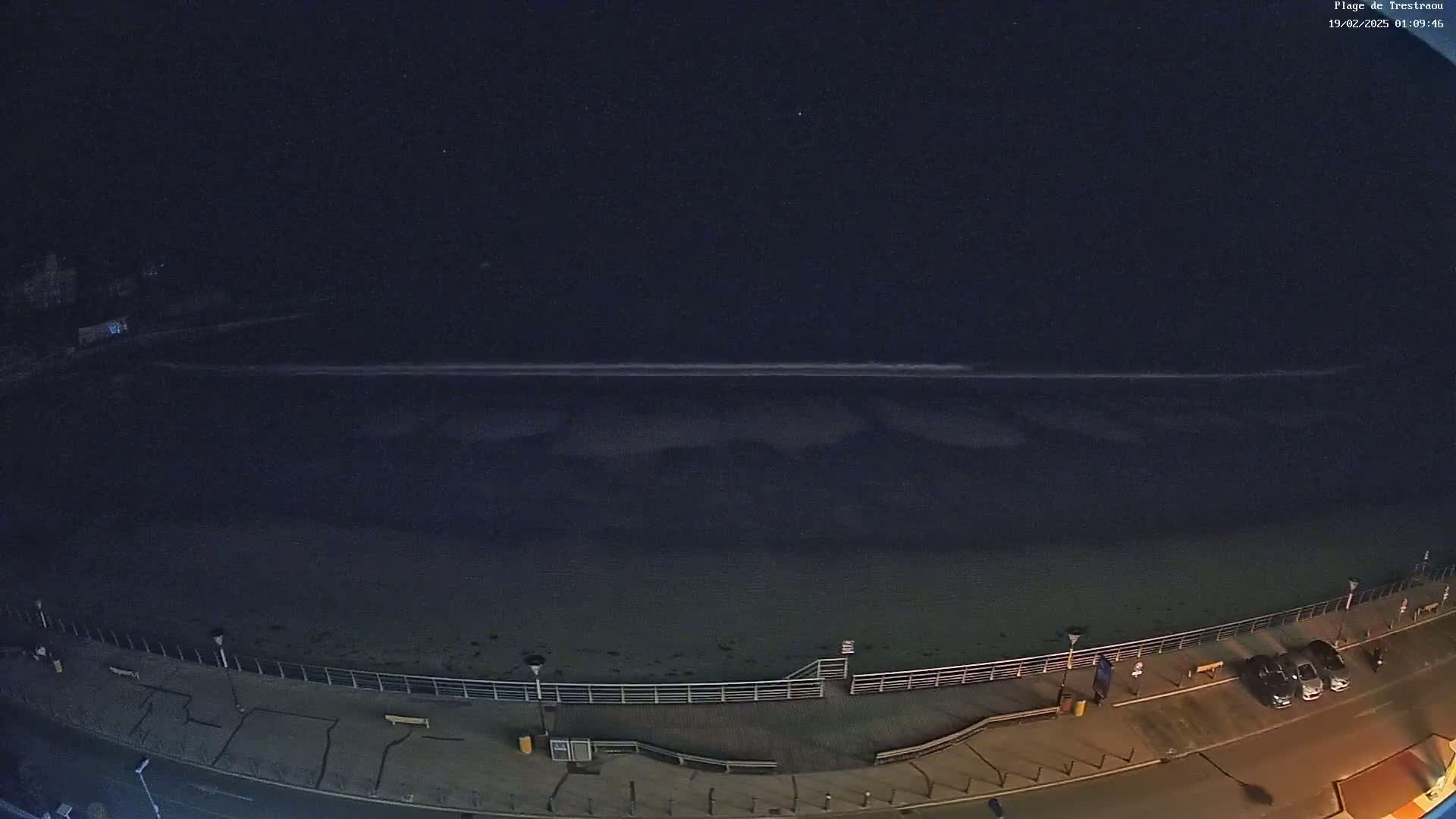 A dark, nighttime view of a beach with calm waves, a boardwalk, and a few parked cars along a road.