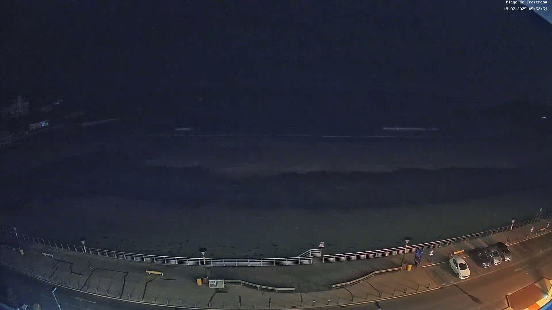 A dark, nighttime beach scene shows a calm ocean under a clear, starlit sky, with a few cars parked along a paved walkway beside a railing.